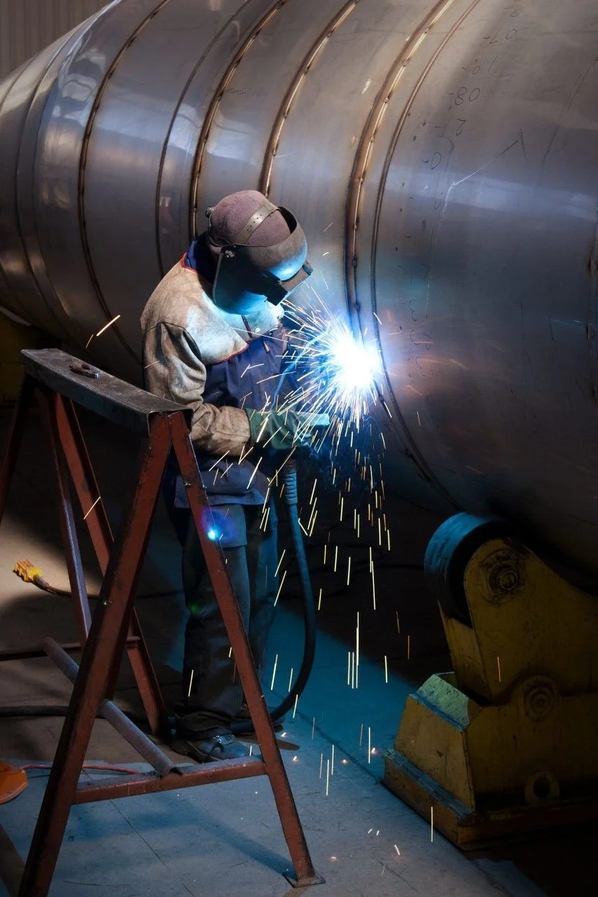 Worker welding a large metal pipe while standing on a ladder in an industrial setting.