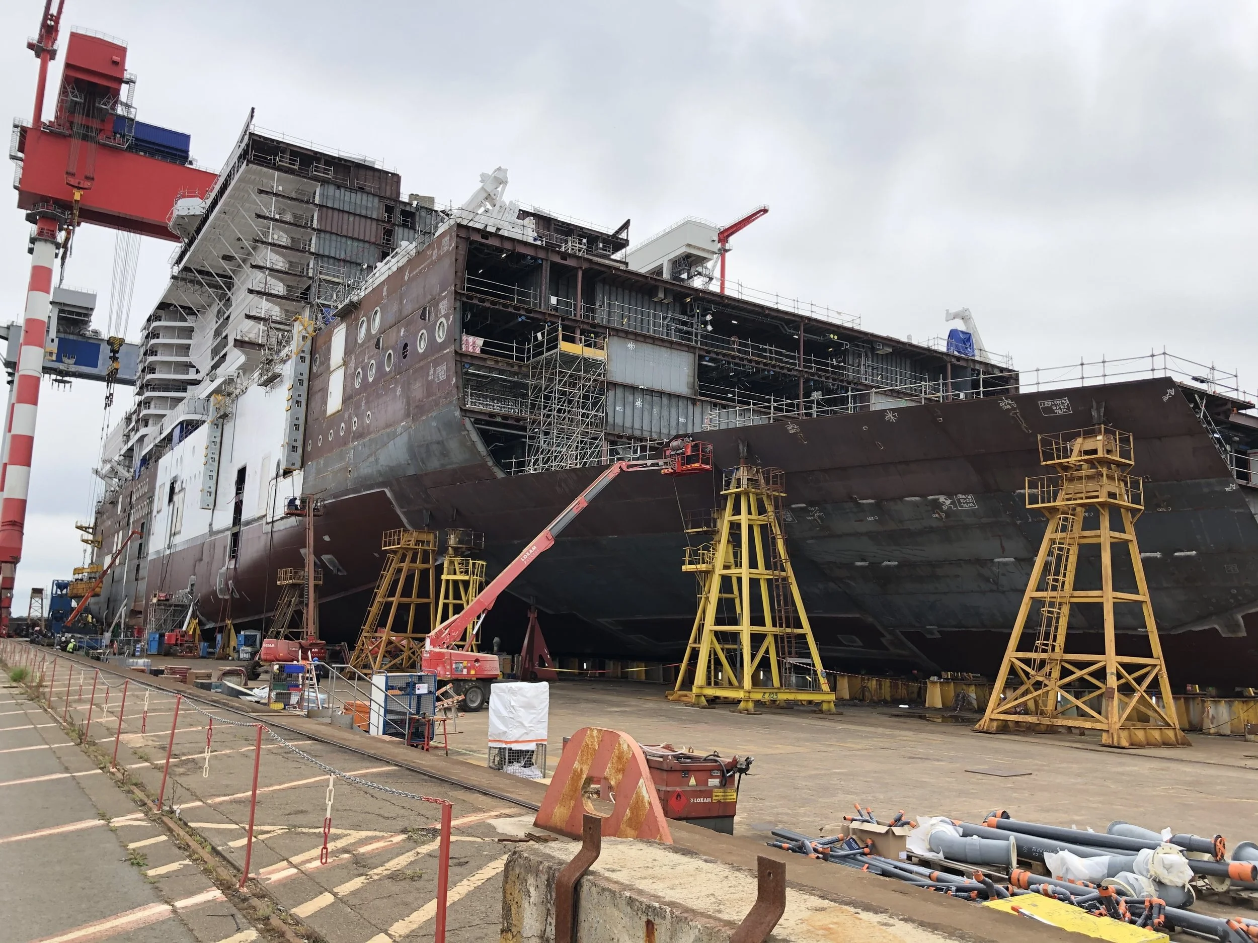 A large ship under construction being held in place by yellow supports, with cranes and construction equipment around it at a shipyard.