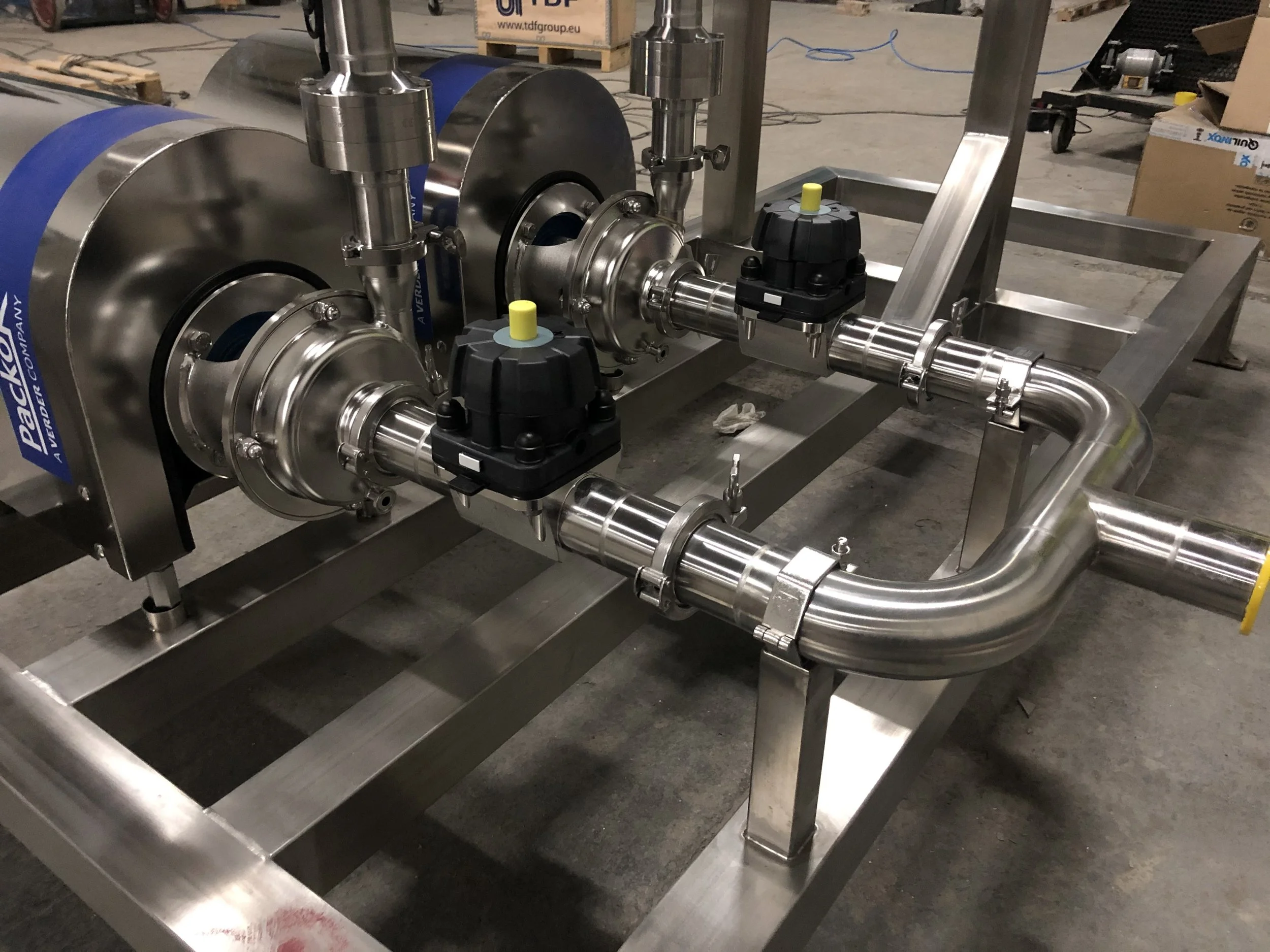 Stainless steel industrial piping system with valves and clamps, mounted on a metal frame, in a warehouse setting with cardboard boxes and equipment in the background.