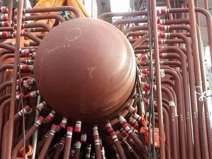 A large bronze or copper sphere surrounded by numerous red and white pipes and valves, possibly part of industrial equipment or machinery.