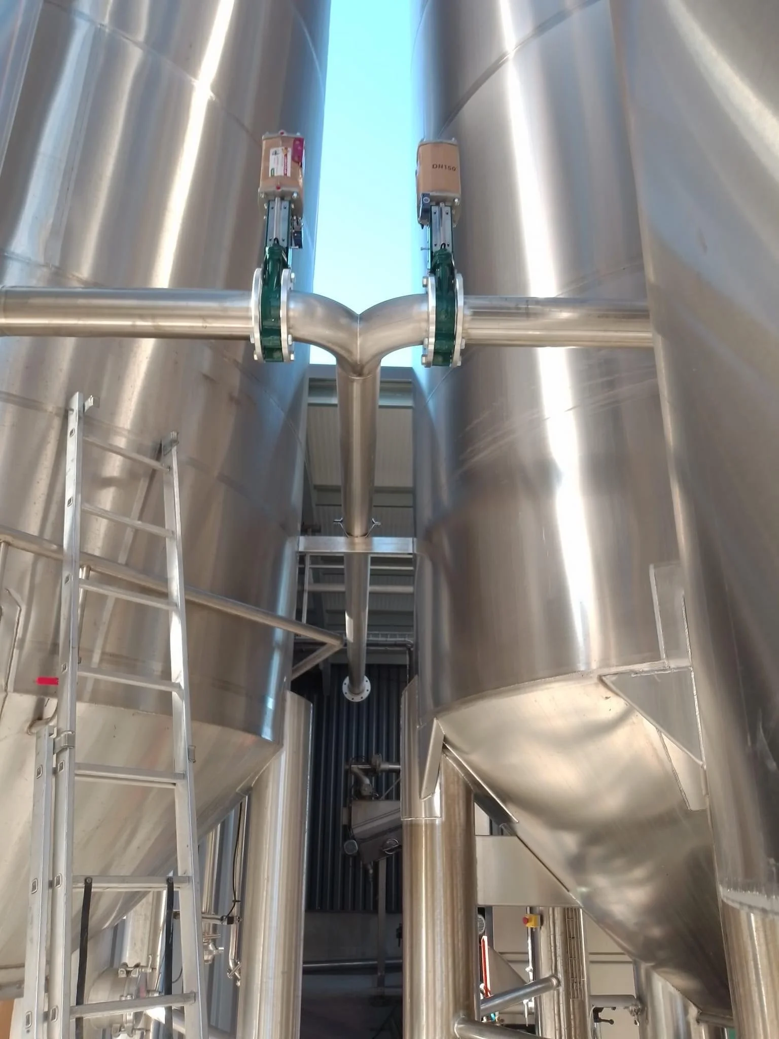 Close-up view of large stainless steel industrial tanks connected by pipes with a ladder on the side in a factory setting.