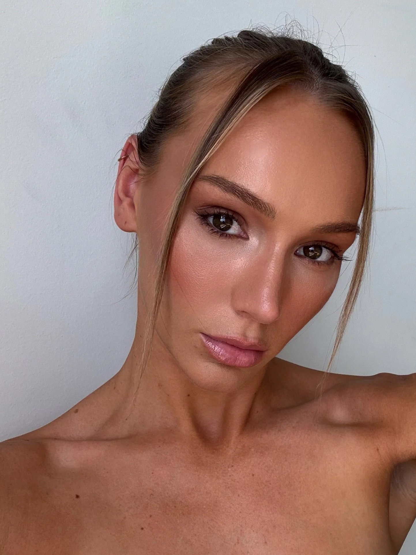 A close-up selfie of a woman with glowing skin, tanned skin, brown eyes, and light brown hair styled slick back, standing against a plain white background.