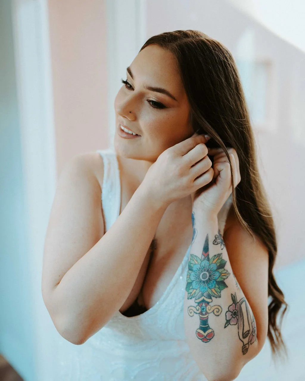 Bride with long brown hair and tattoos is putting on earrings, smiling softly, wearing a white sleeveless top, in a bright room. getting ready in her bridal suite. glowy warm weddings.