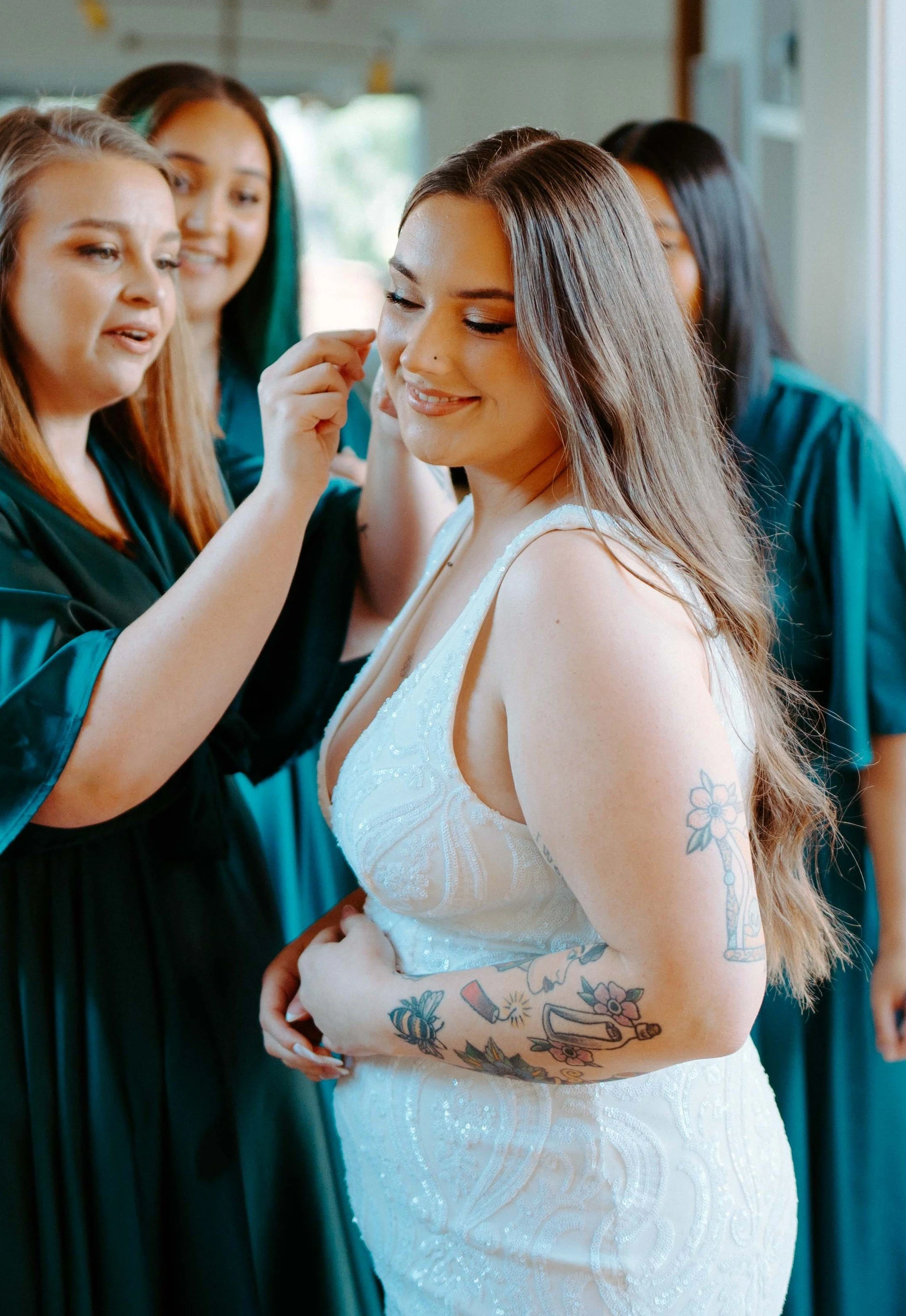 A woman in a white dress with tattoos on her arm is having makeup applied by a makeup artist while other women in matching dark green robes watch, in a brightly lit room.
