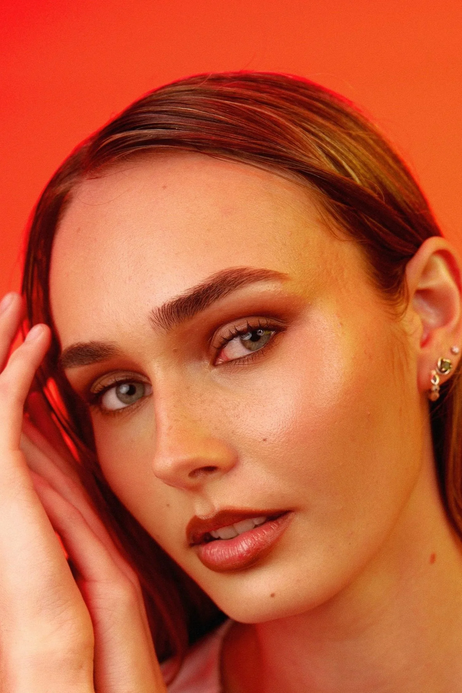 Close-up of a woman with brown hair, blue eyes, and full lips, posing with her hand near her face against an orange background.