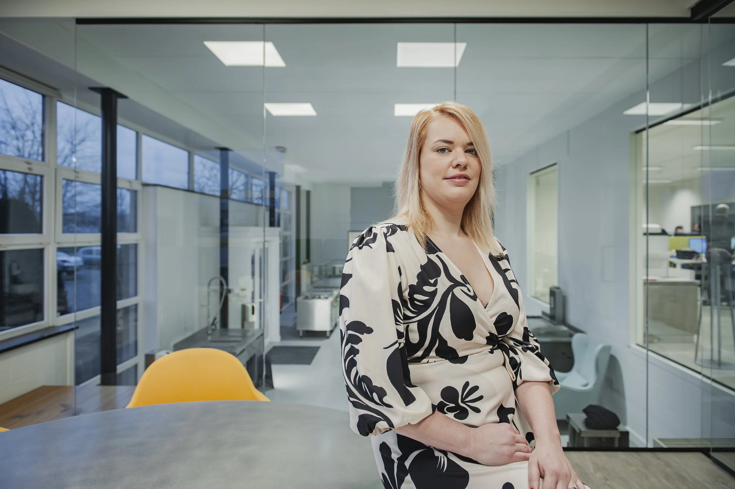 Een vrouw staat in een modern kantoor, met grote ramen en een glazen scheidingswand, kleedt in een zwart-wandelwit bloemenprint jurk.