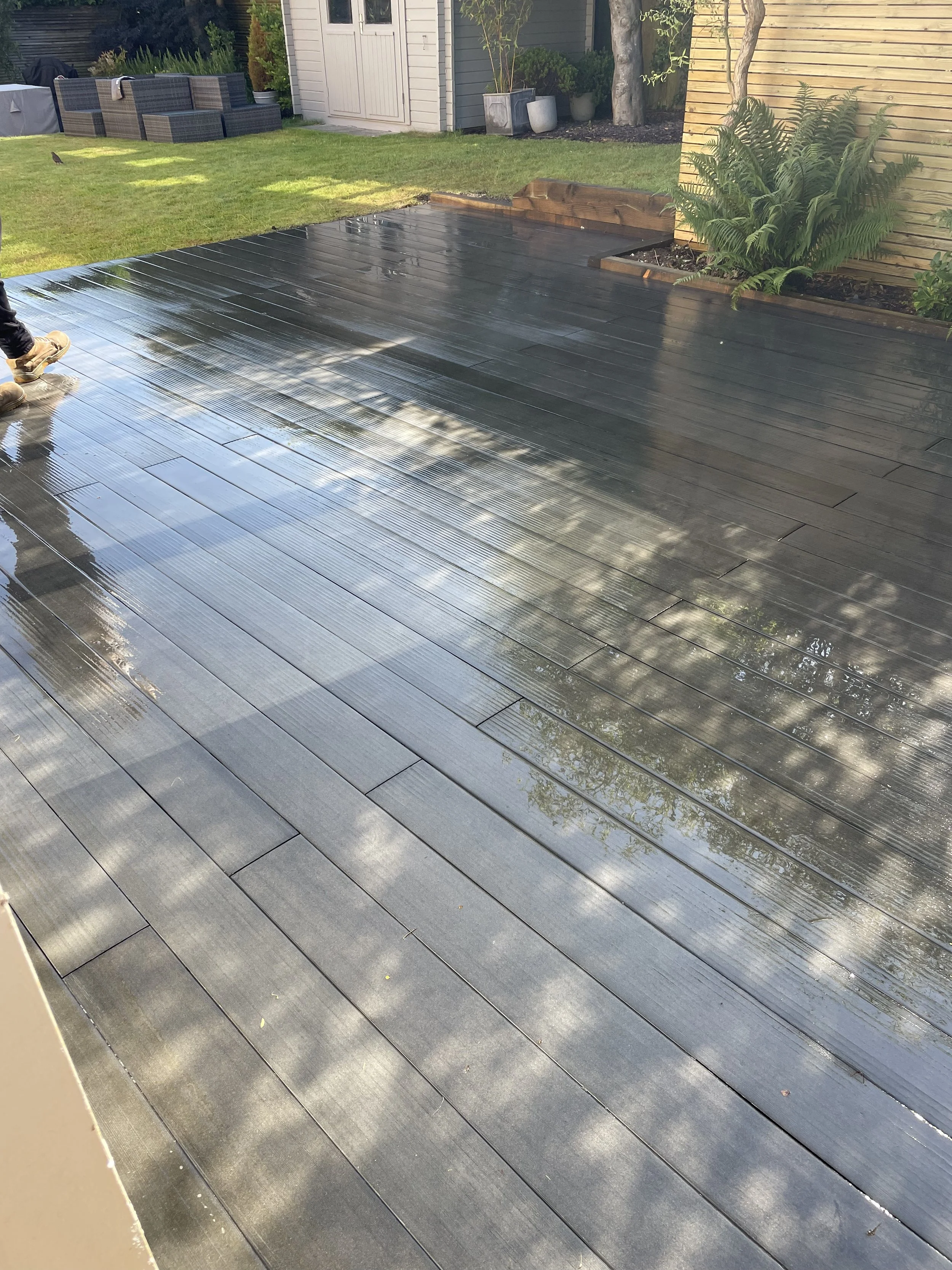 A newly waterproofed wooden deck with recently applied shiny black waterproofing coating, adjacent to a grassy yard with patio furniture and plants.
