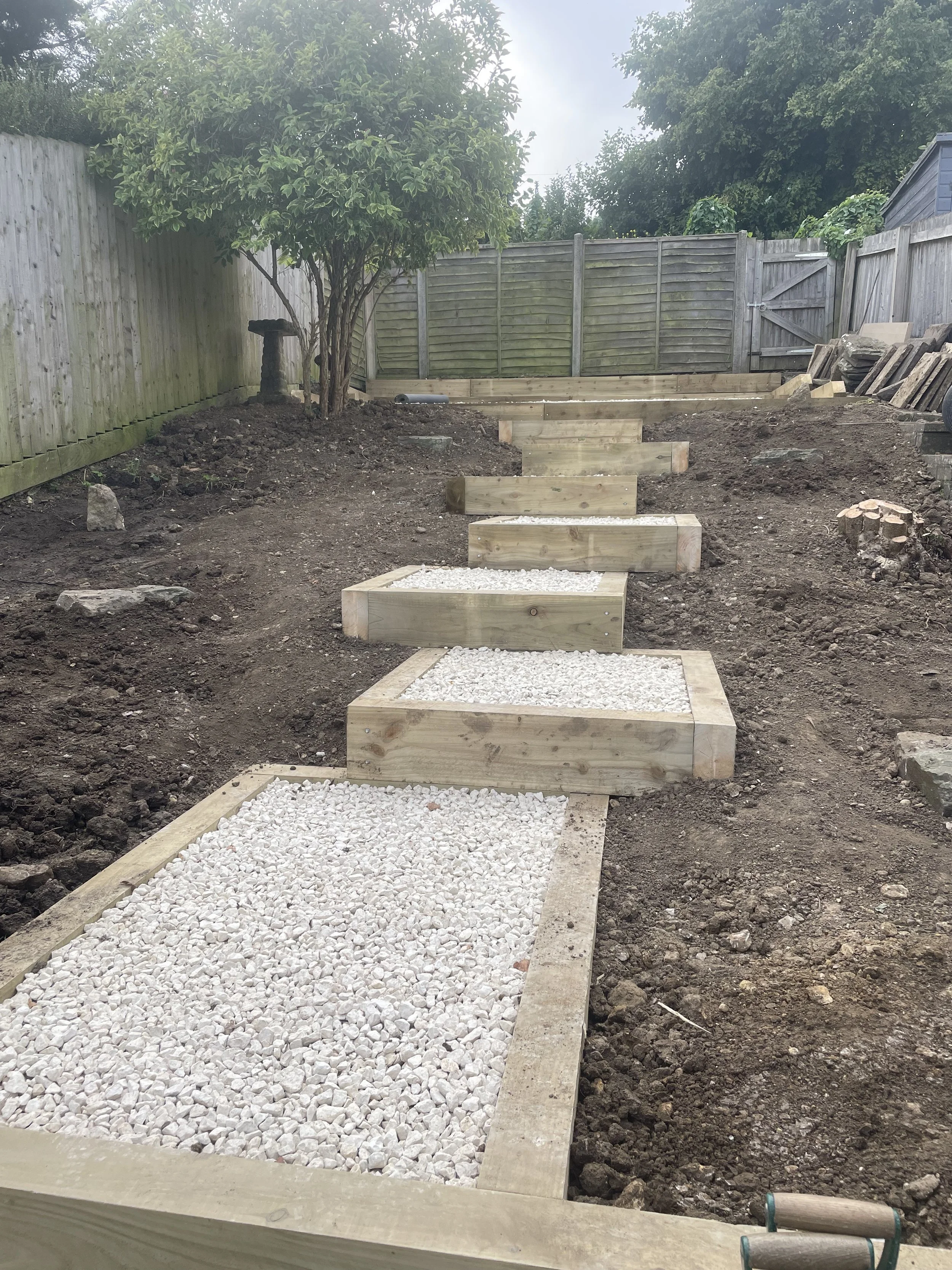 A backyard garden pathway being constructed with wooden frames filled with white gravel on a sloped ground. The pathway includes a series of rectangular steps, also filled with white gravel, leading towards a fenced area with trees and a blue shed in the background.
