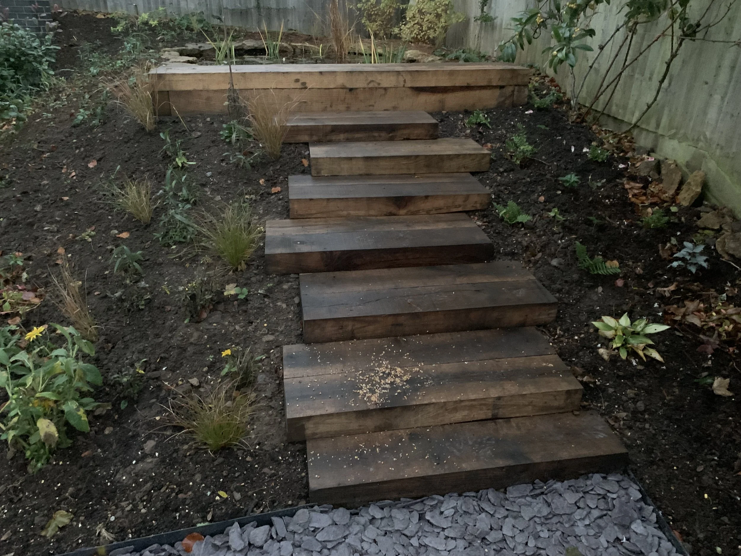 Wooden outdoor staircase leading to a raised garden bed with plants, surrounded by dark soil, small plants, and decorative rocks along the edge.