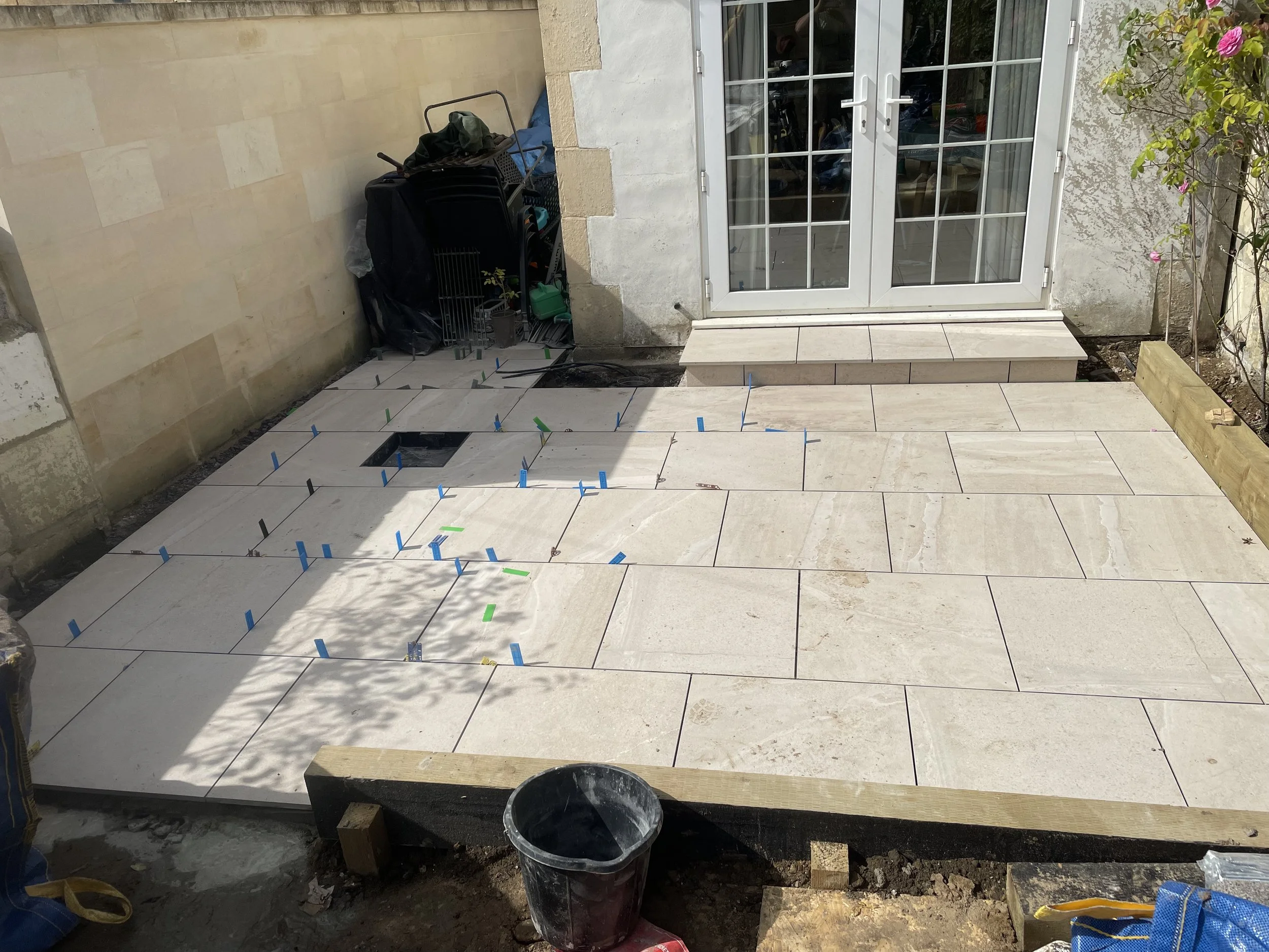 Exterior patio area under construction with tiles being laid, and a white double glass door leading inside. Tools and materials are visible around the area.