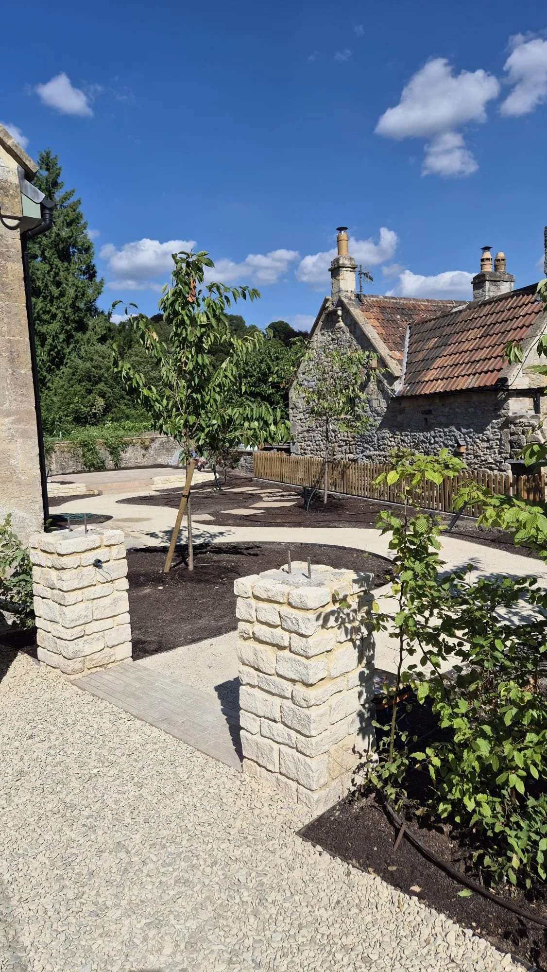 A newly landscaped yard with young trees, stone pillars, and gravel walkways under a blue sky with clouds, surrounded by old stone houses with tiled rooftops.