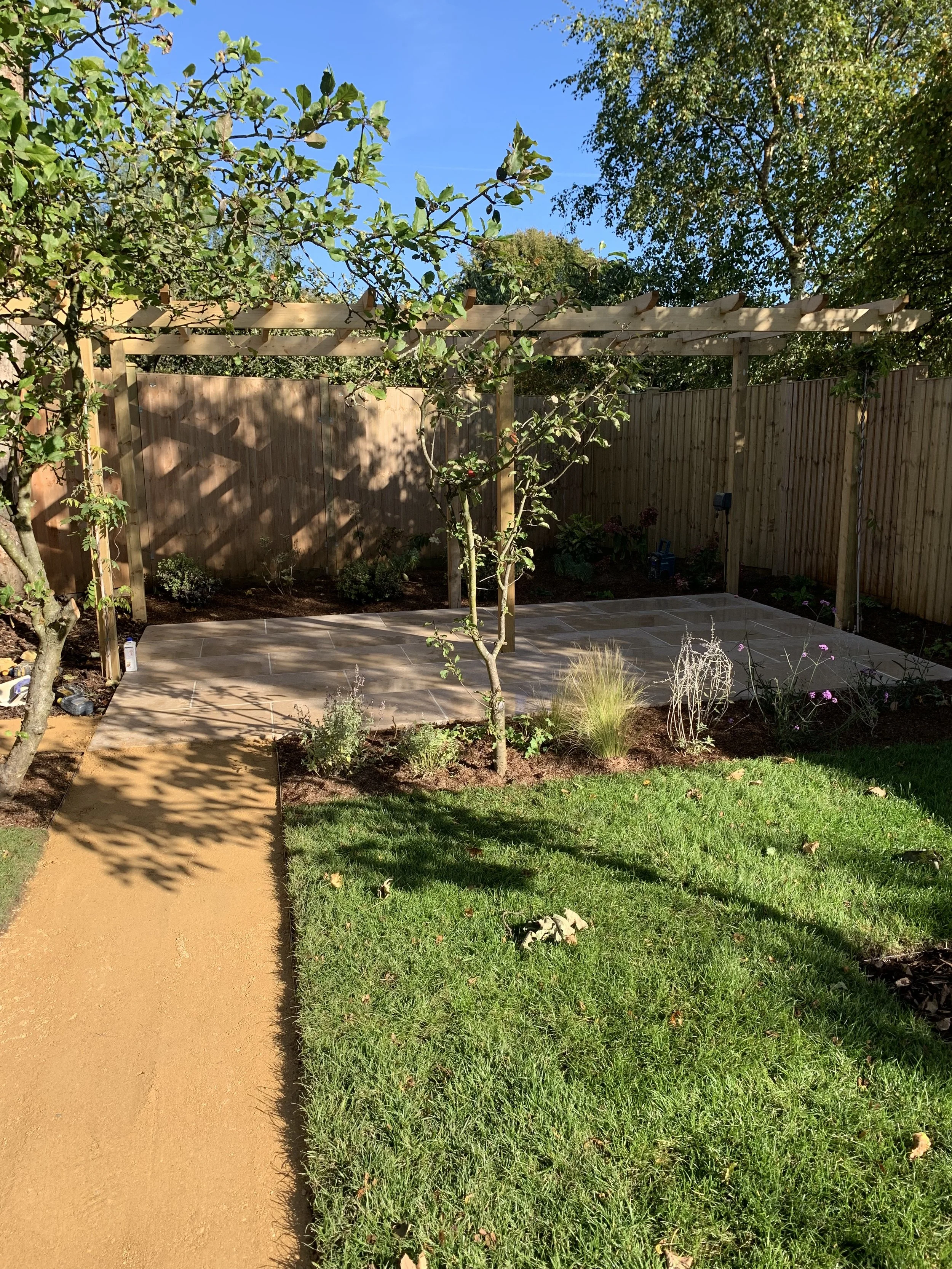 View of a backyard garden with a small tree, flower bed, pathway, and a wooden pergola frame with shadows cast on the ground, surrounded by a wooden fence and green grass.