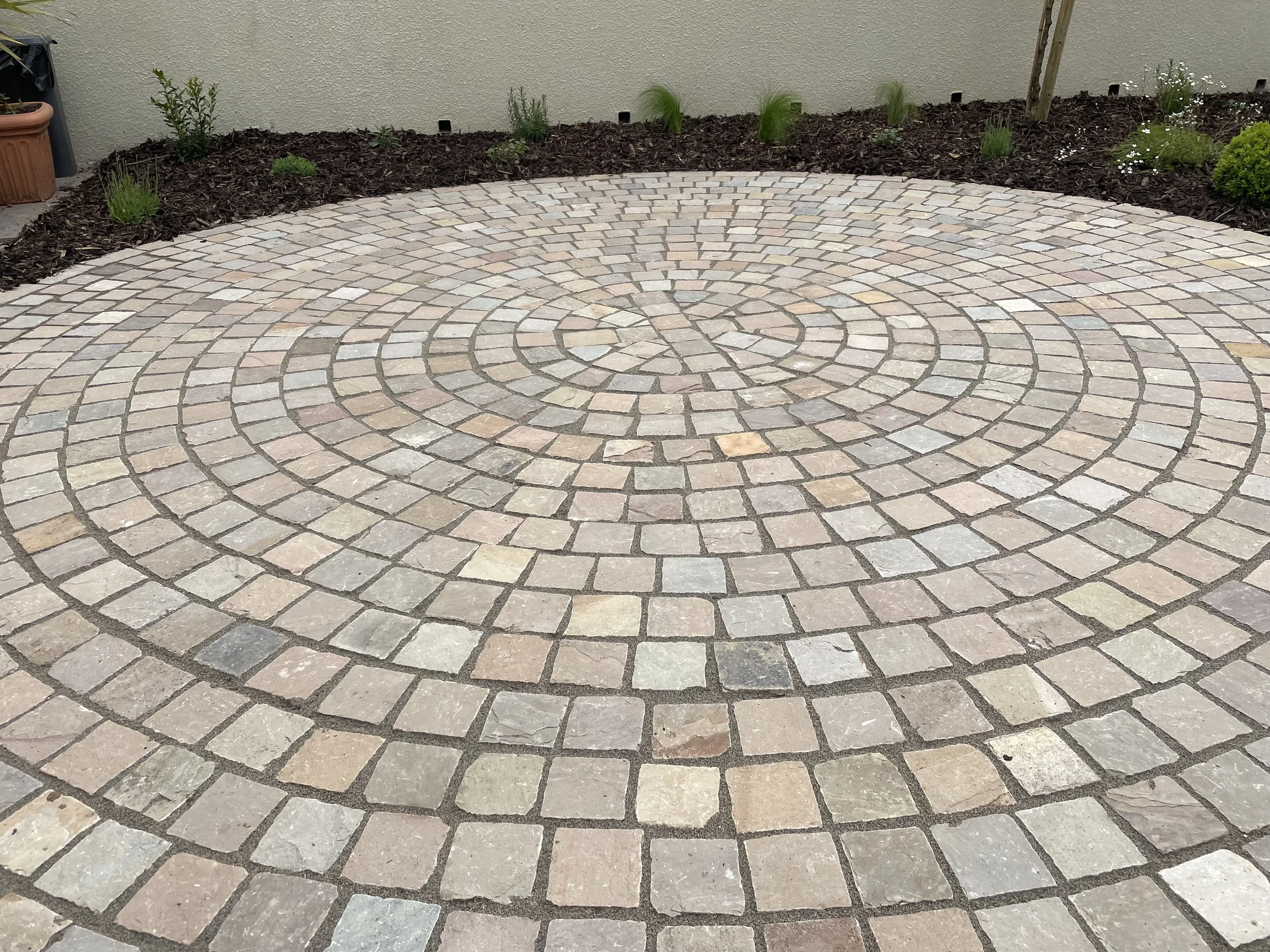 A circular pattern of multicolored stone pavers on a patio, bordered by a garden bed with small plants and mulch.