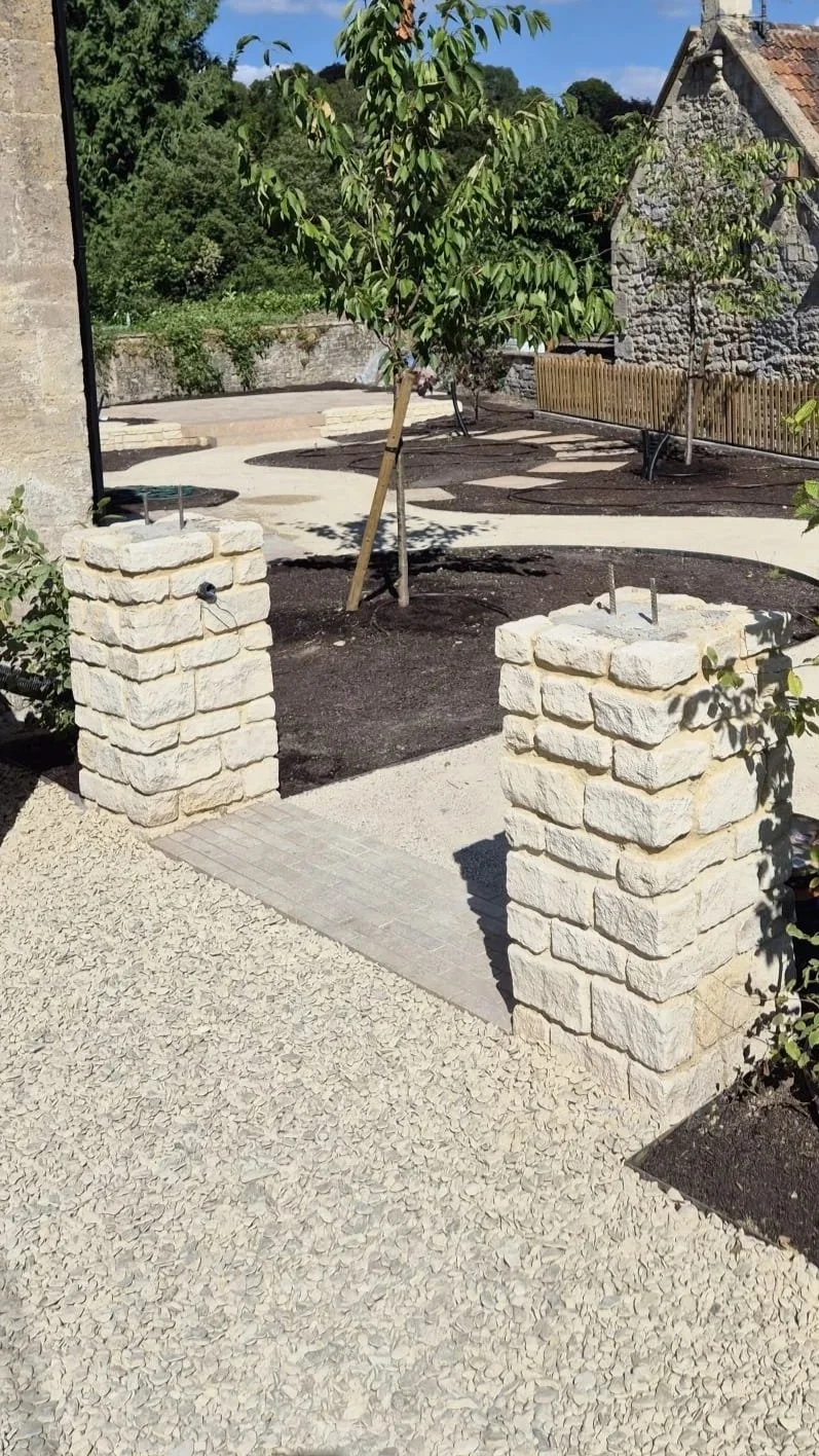 A garden with young trees, stone pillars, gravel, and a winding paved pathway, with a stone house and dense greenery in the background.