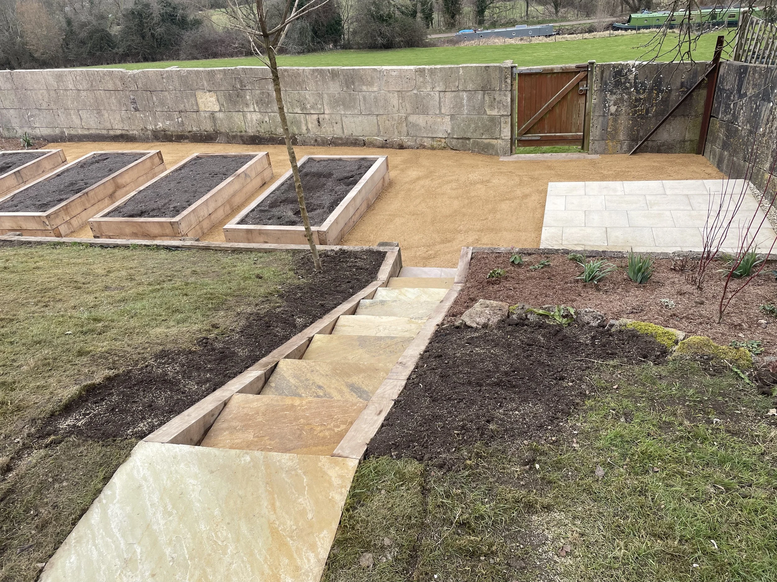 A backyard garden with wooden planter boxes, a small tree, and a paved patio area, with a stone wall and a green lawn in the background.