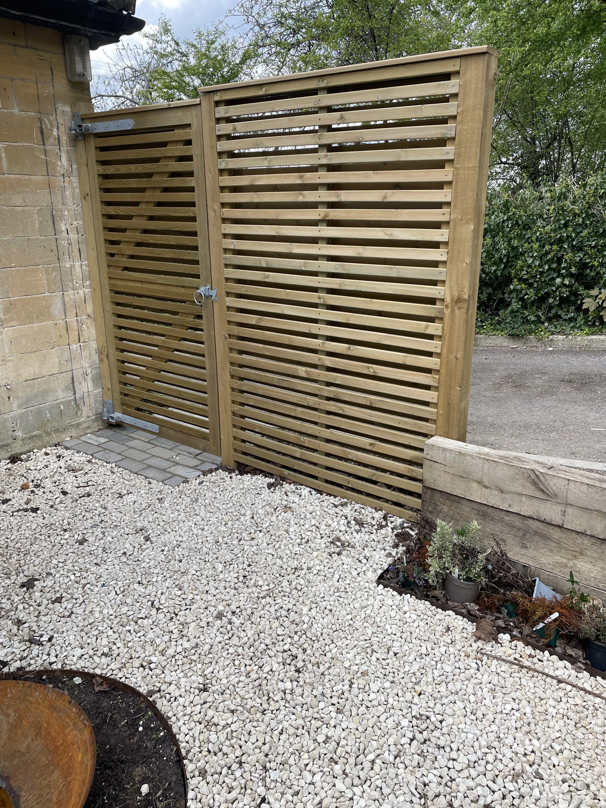 A newly built wooden gate with horizontal slats and a lock, attached to a brick wall on the left side, with gravel ground and some small garden plants on the right side.
