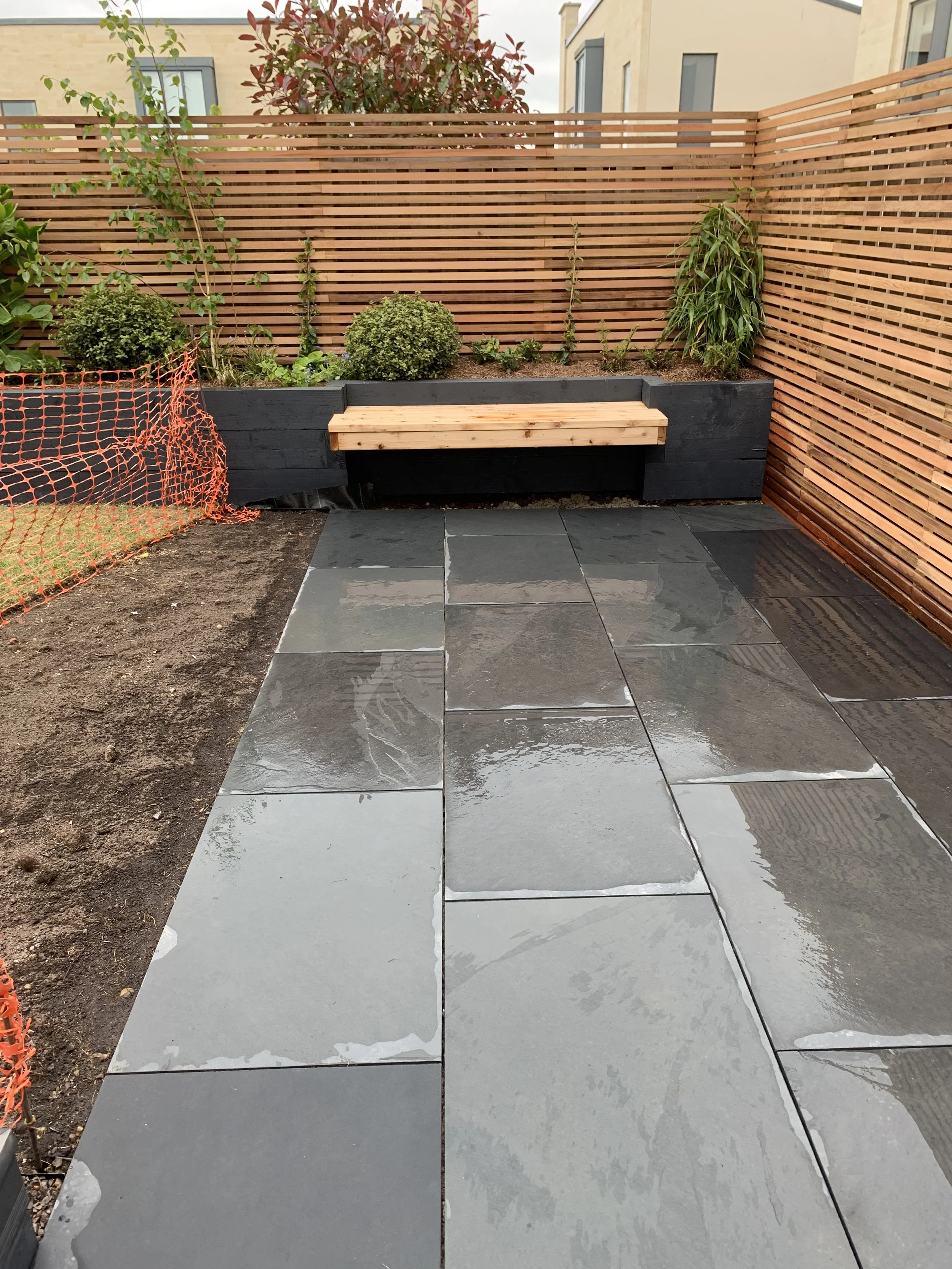 A modern outdoor patio area with wet, shiny gray tiles, a wooden bench, and a raised garden bed with plants and shrubs, enclosed by wooden slat fencing.