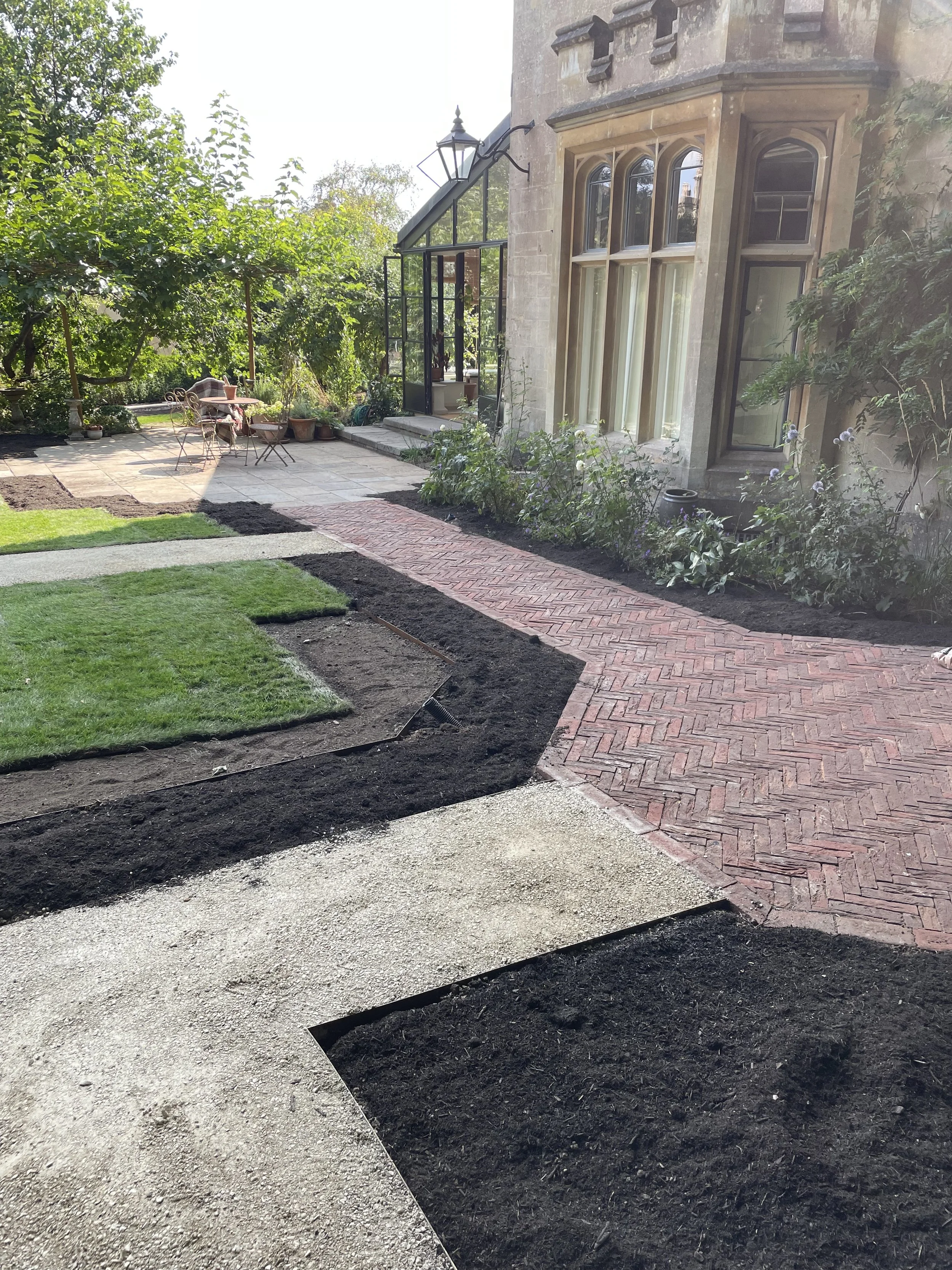 A garden with a brick pathway, patches of green grass, and freshly tilled soil, adjacent to a historic stone building with large windows and a glass conservatory.