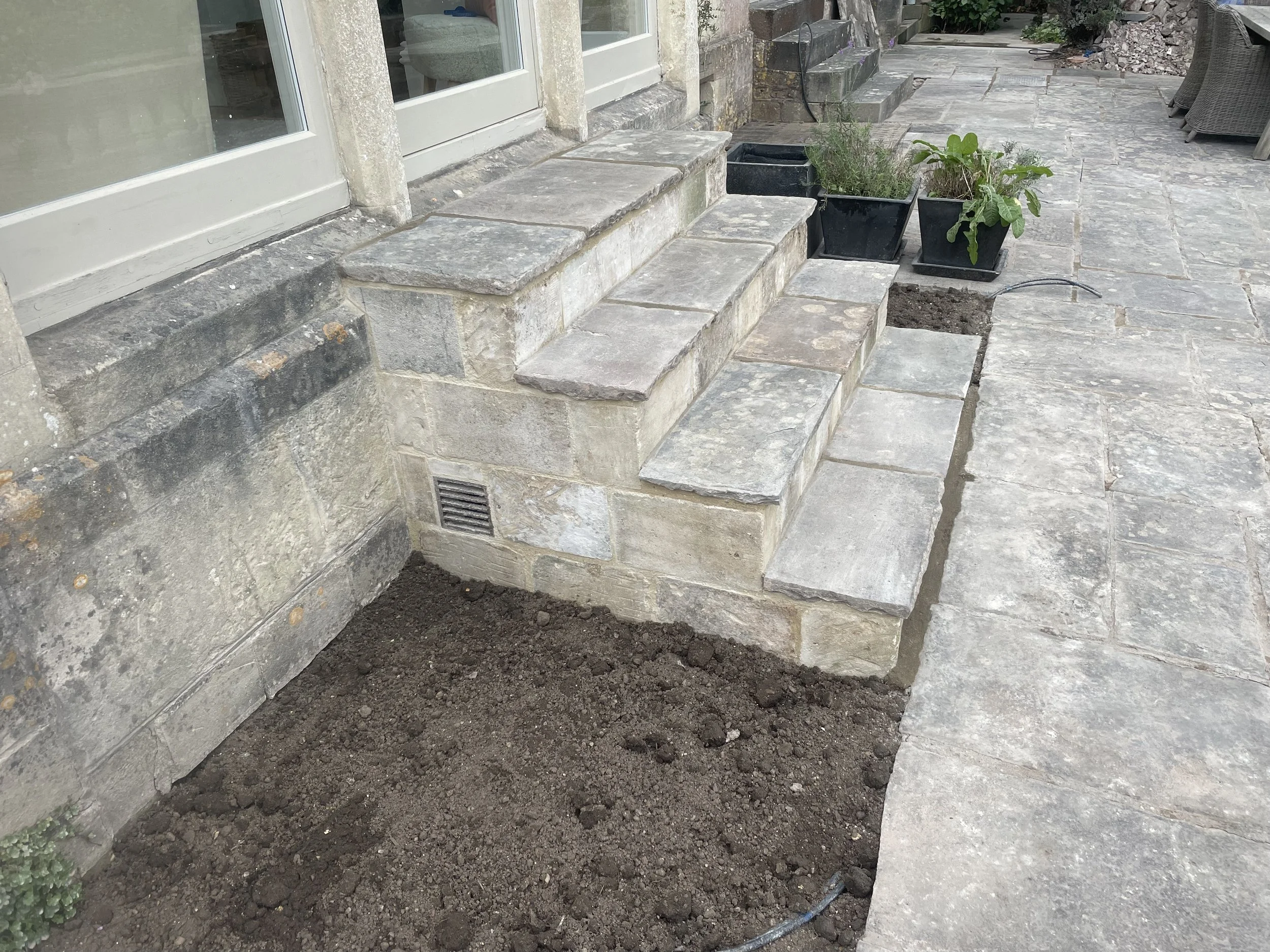 Stone patio and steps outside a house, with flowerpots and a small patch of soil.