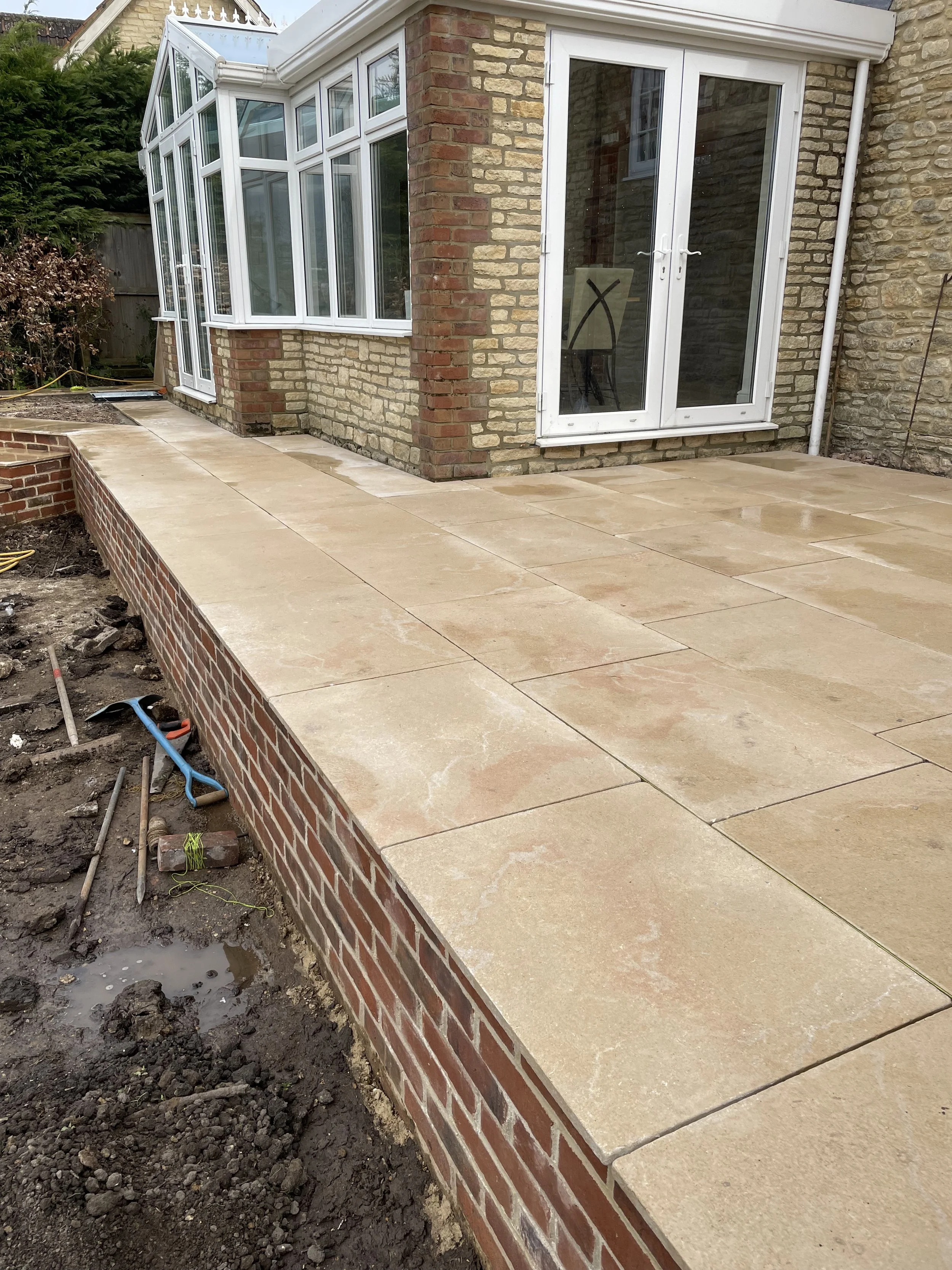 A newly constructed patio with beige stone tiles and a brick retaining wall, attached to a brick house with white-framed windows and a glass door, with construction tools nearby.