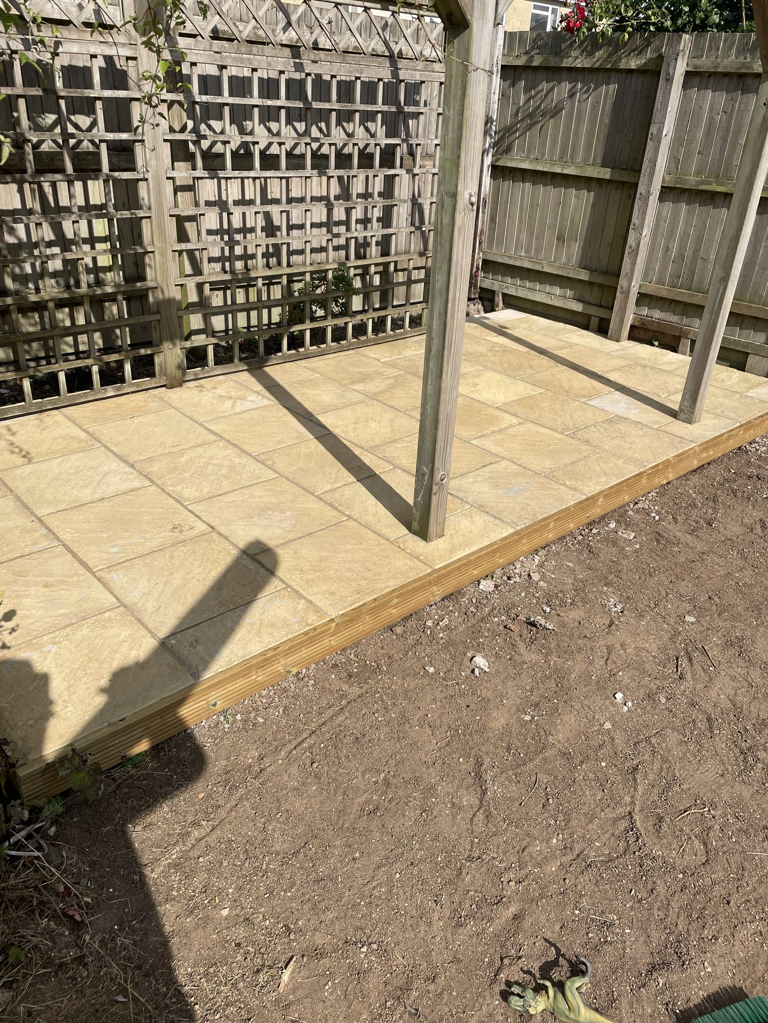 A small wooden deck with a tiled surface, surrounded by a garden fence, with a garden glove and a garden tool on the ground nearby.
