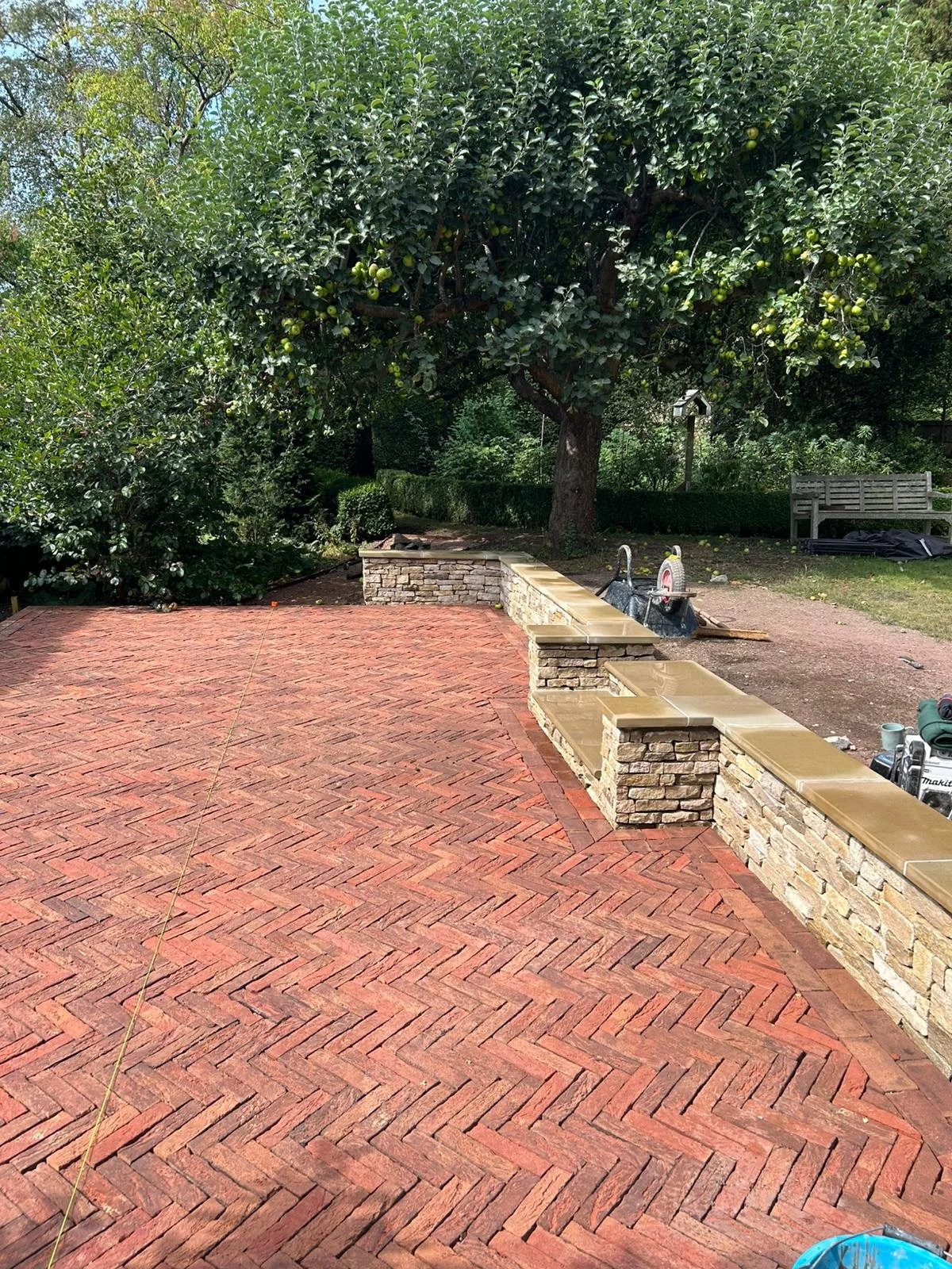 A newly constructed brick patio with a stone and brick retaining wall, a large tree, and garden furniture in the background.