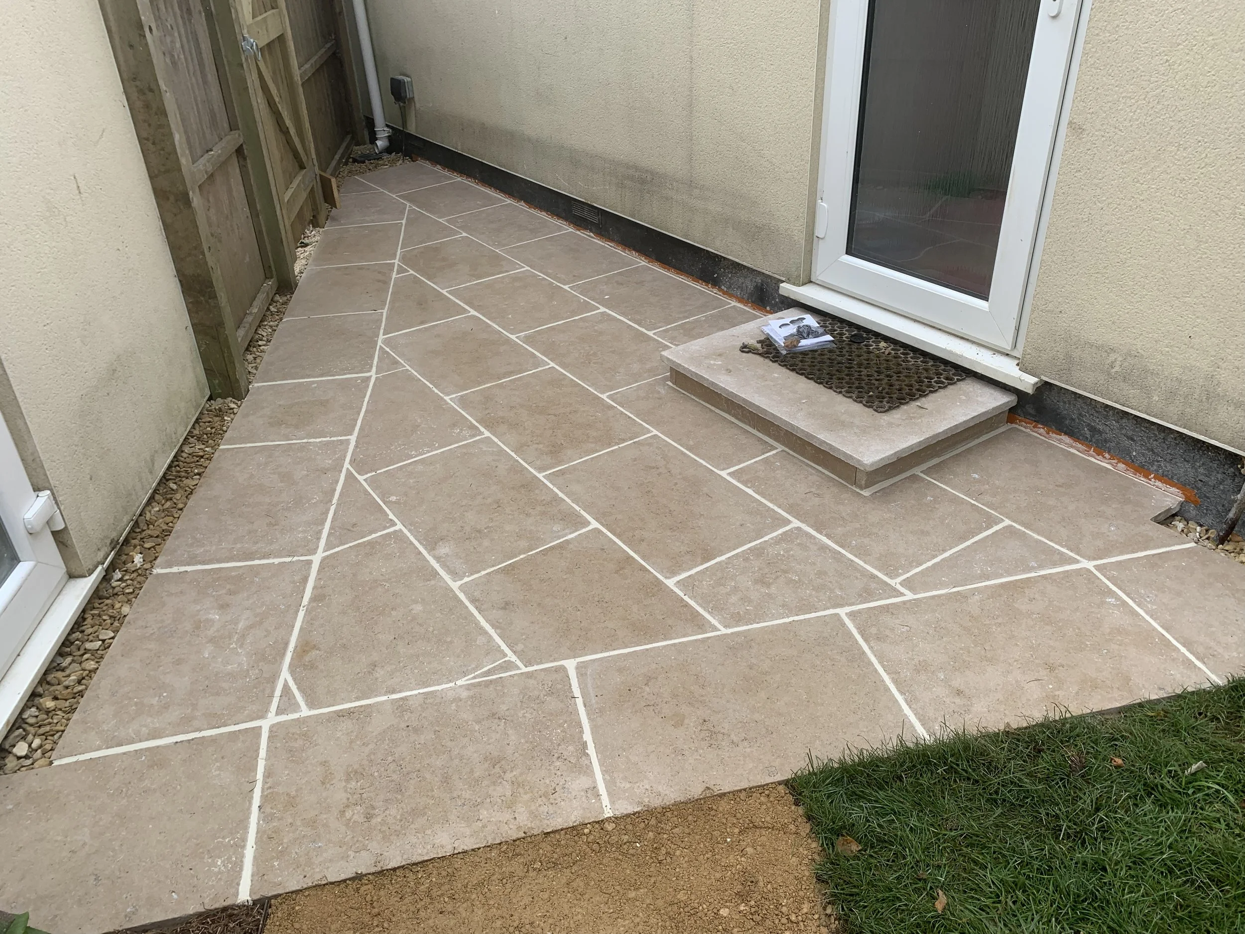 A newly constructed stone patio with beige tiles in an outdoor space, adjacent to a house with a sliding glass door, a doormat, and a small raised concrete step.