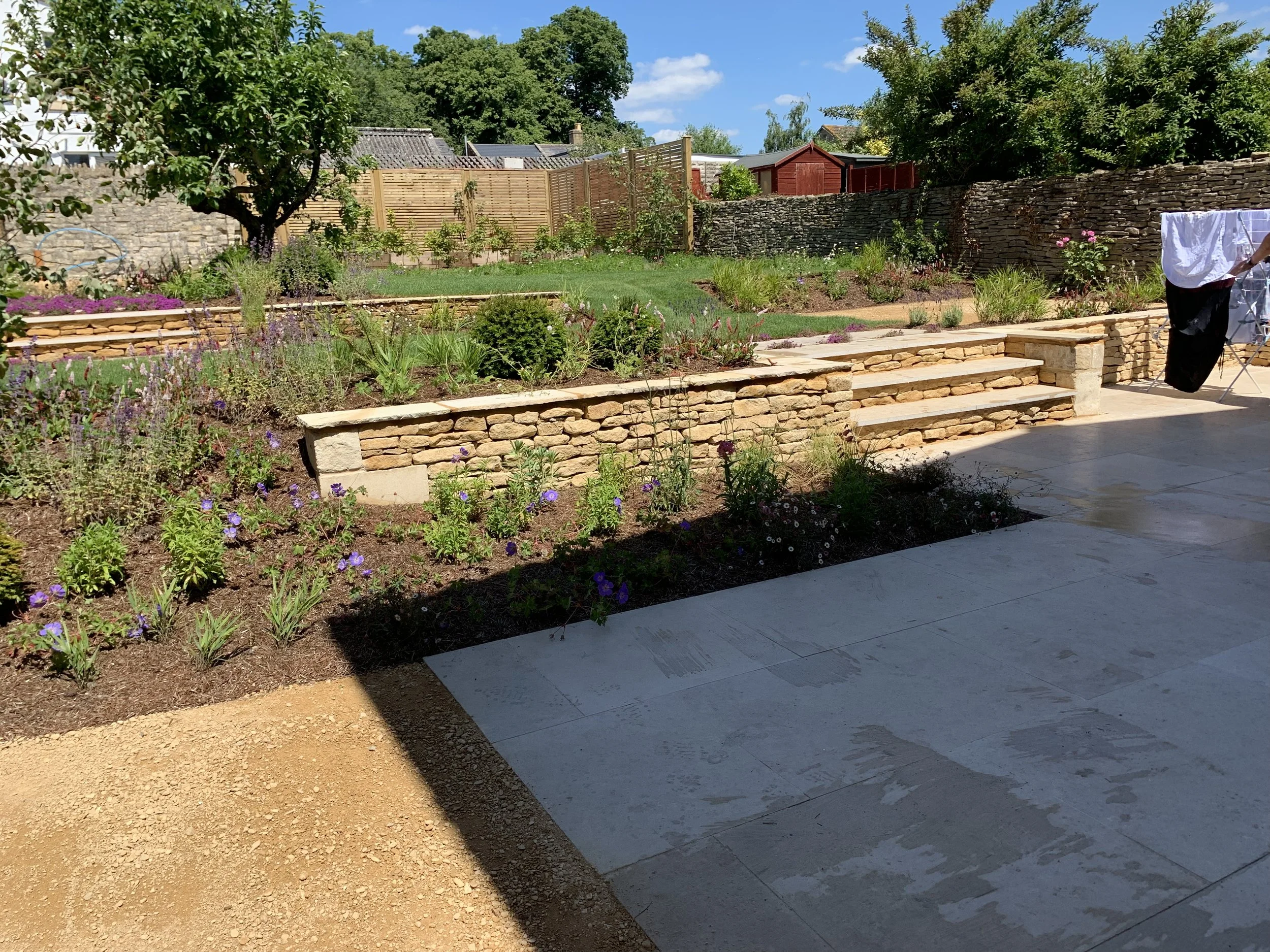 A landscaped backyard garden with trees, shrubs, and flower beds, featuring a stone retaining wall and steps, a patio area with hanging laundry, and a sunny blue sky.