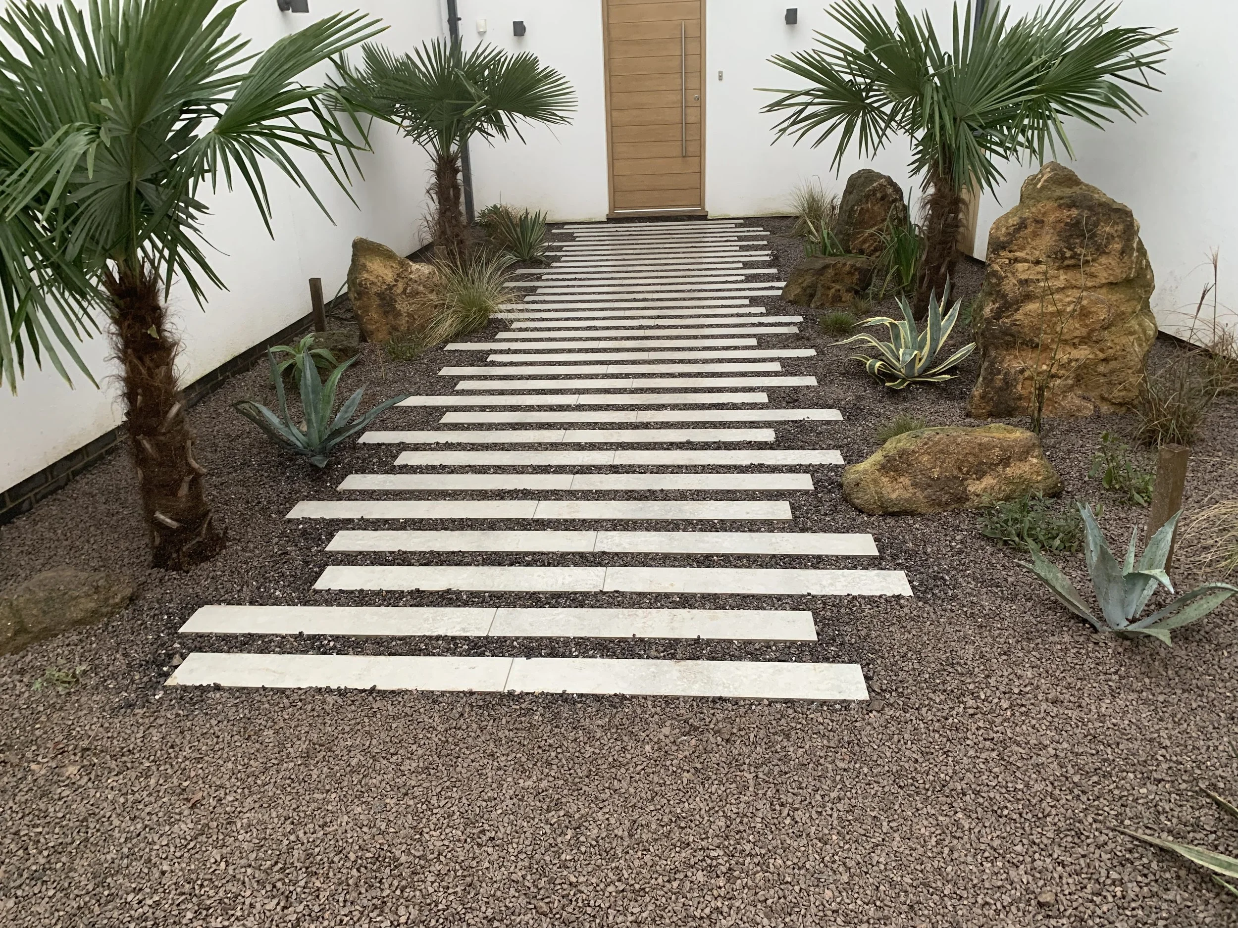 A modern pathway with white rectangular stepping stones bordered by gravel, surrounded by a desert-themed garden with palm trees, agave plants, and large rocks, leading to a wooden door.