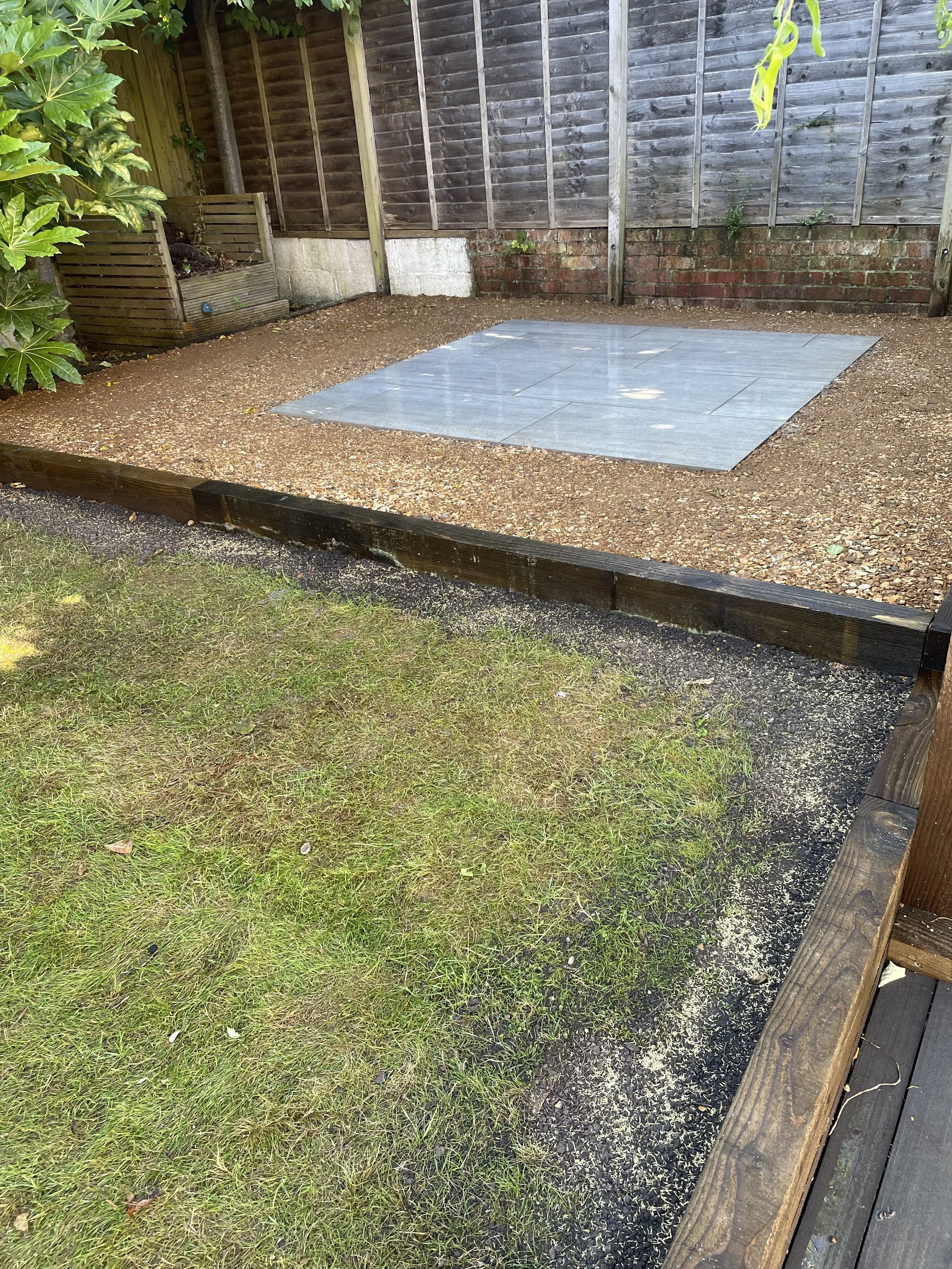 A backyard patio area with a section paved with light blue tiles, surrounded by gravel and grass, enclosed by wooden fencing and a brick wall.