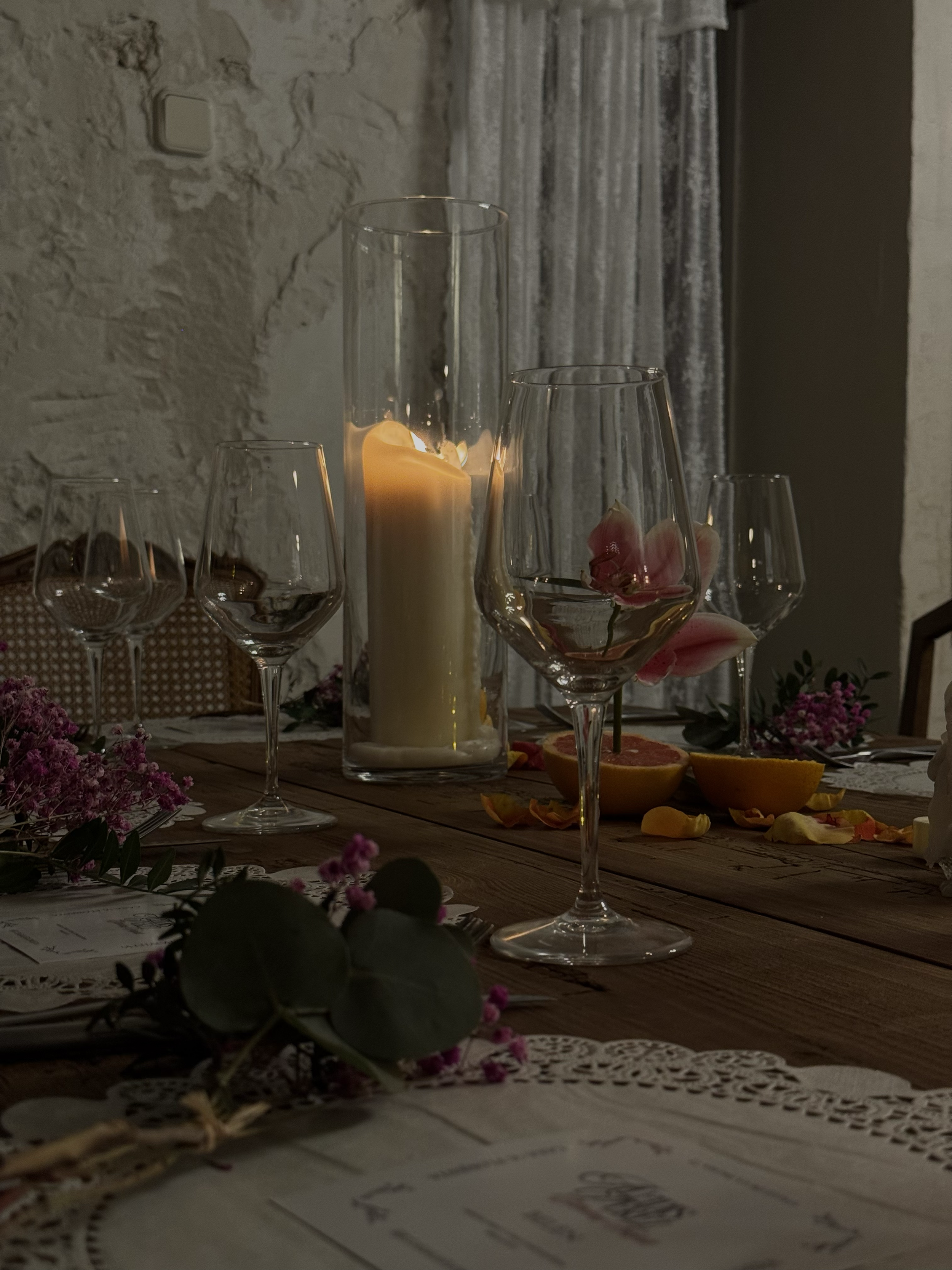 Mesa decorada con un candelabro con vela encendida, varias copas de vino, flores, y rodajas de cítricos, en un ambiente rústico y acogedor.