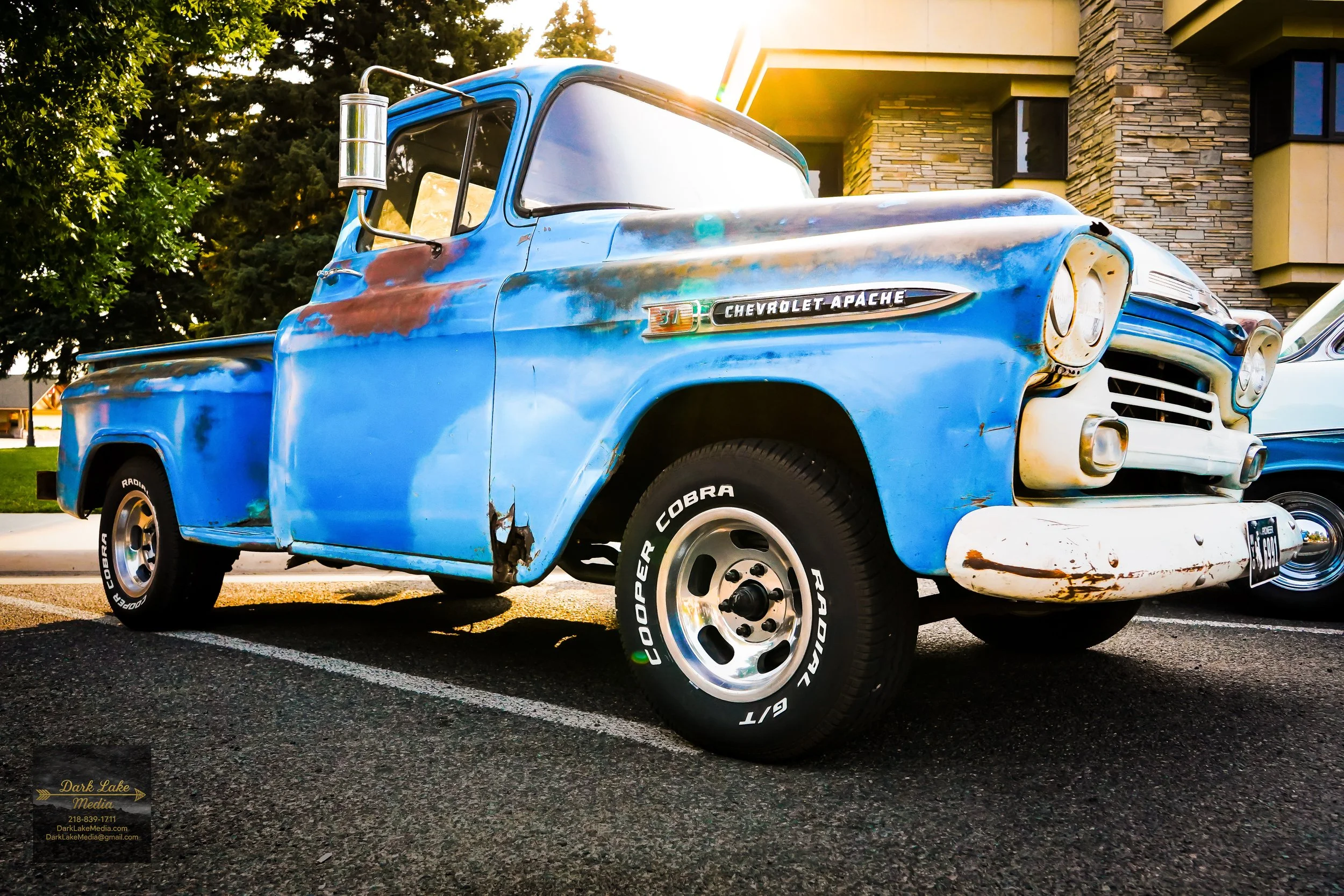 An old blue Chevrolet Apache pickup truck with rust spots and damage parked on a street, with a building and trees in the background.