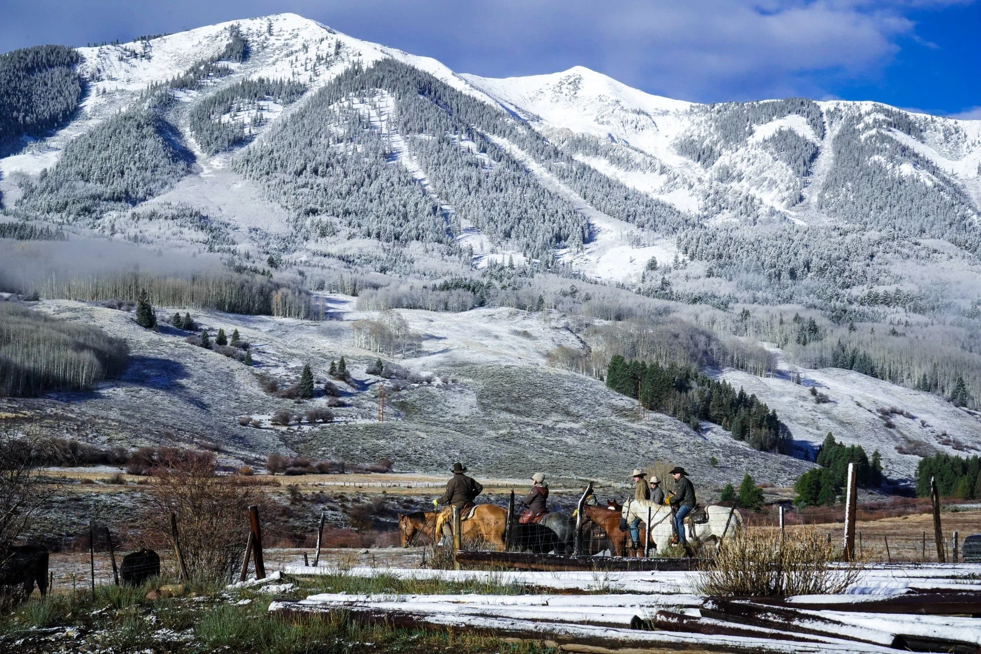 A group of people riding horses on a trail in a snow-covered valley with mountains and evergreen trees in the background.