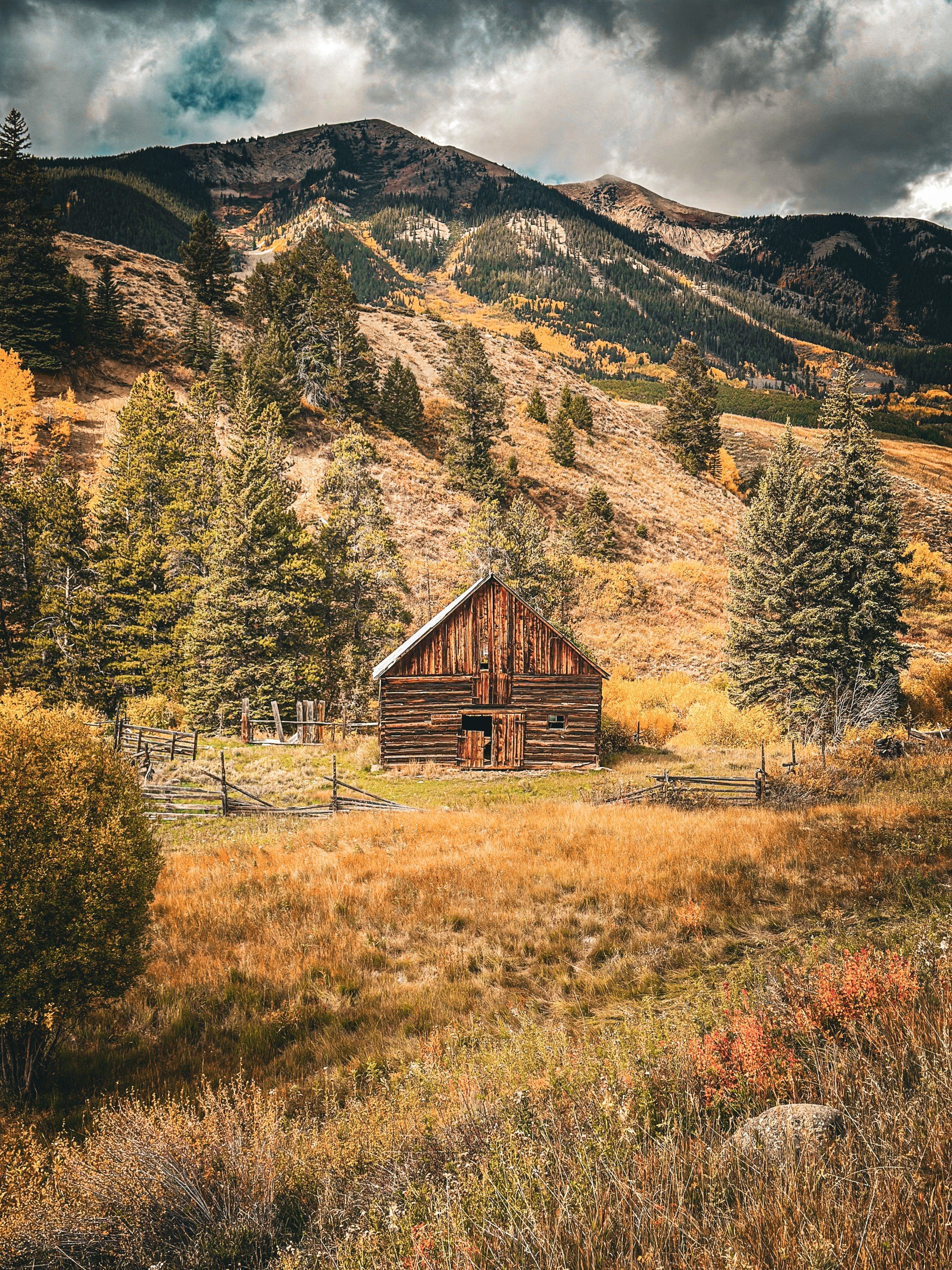 A rustic wooden barn in a fall landscape with mountains, pine trees, and golden foliage.