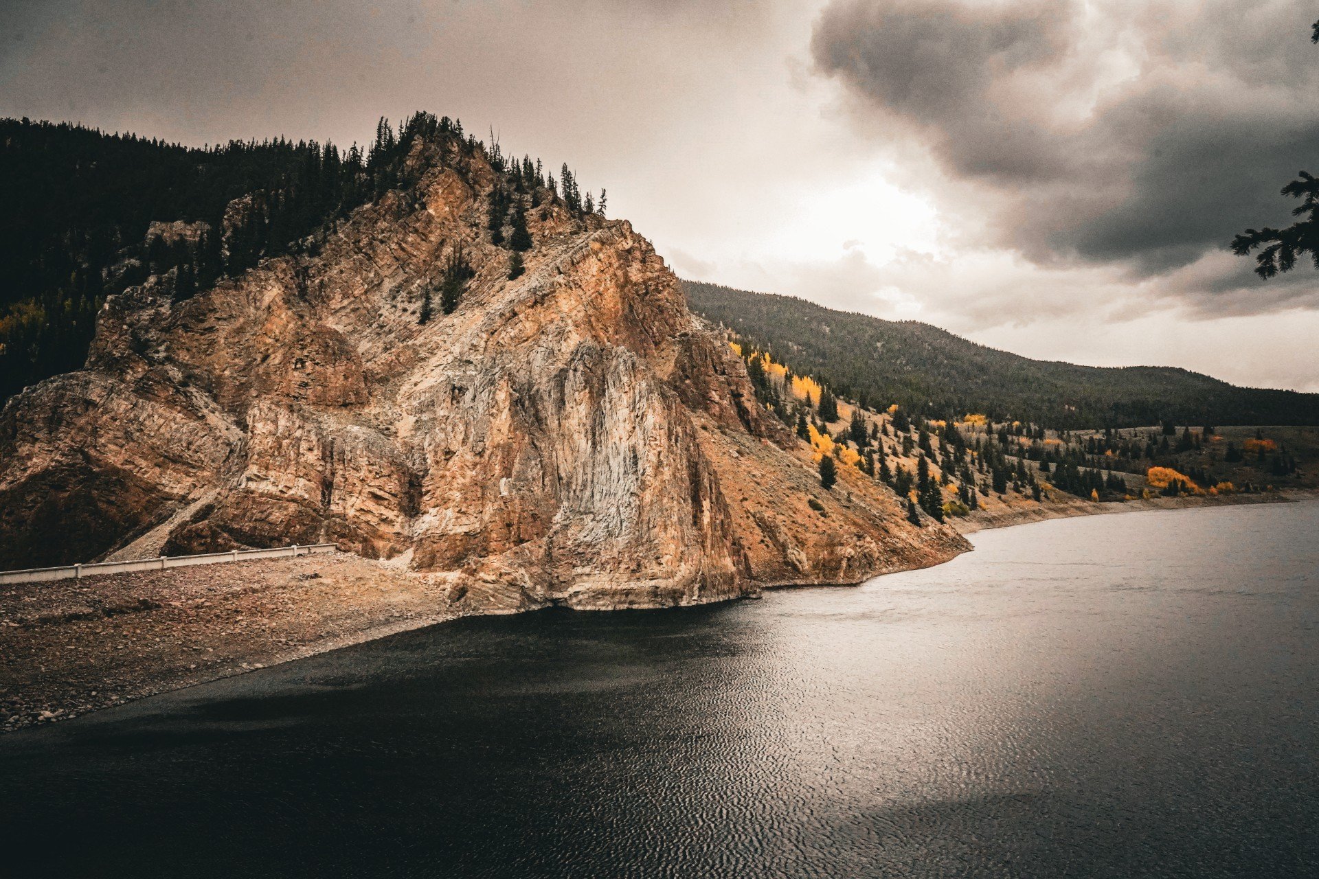 A large rocky mountain with sparse trees, beside a dark body of water under cloudy sky.