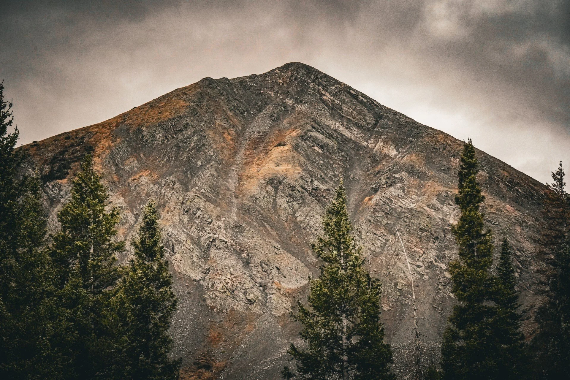 A rugged mountain with visible layers and diagonal formations, surrounded by tall green pine trees, under a cloudy sky.