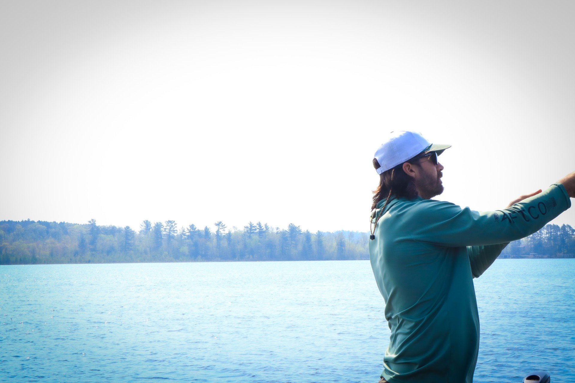 A man with a beard wearing sunglasses, a white cap, and a teal long sleeve shirt standing by a lake.