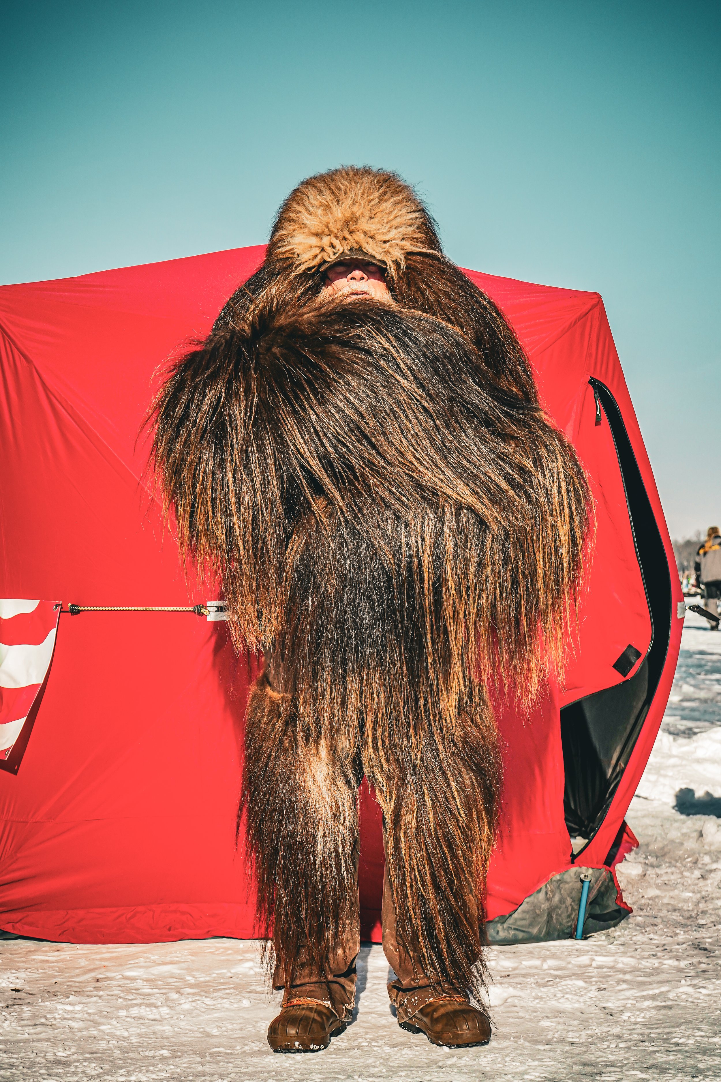 Yeti-like figure with long fur standing in front of a red tent in a snowy landscape.