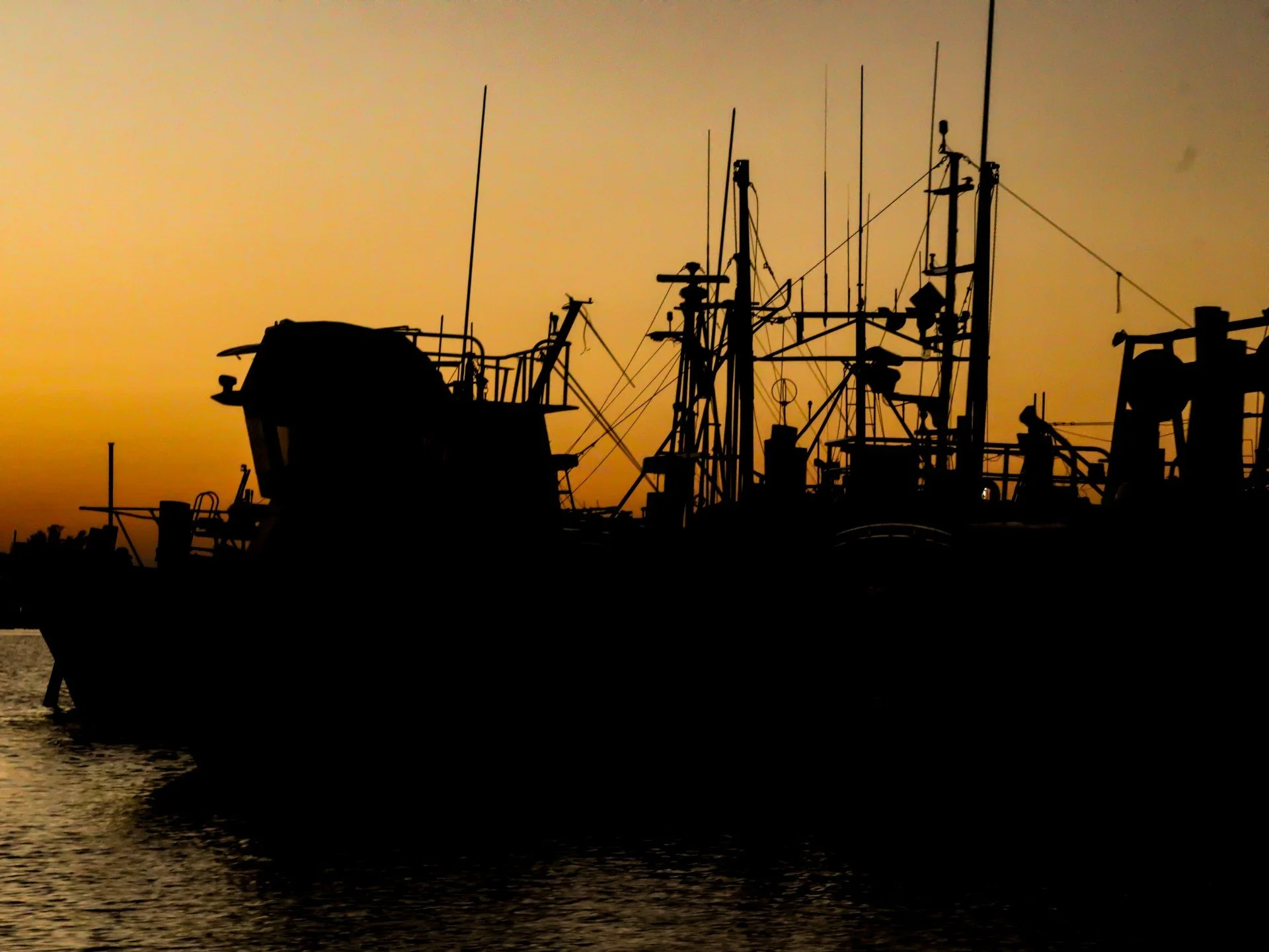Silhouette of boats docked at a harbor during sunset with a yellow-orange sky.