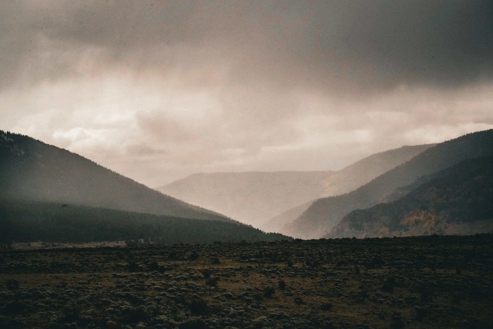 A landscape of a mountain valley with dark, foggy, overcast sky, distant mountains, and a grassy plain in the foreground.