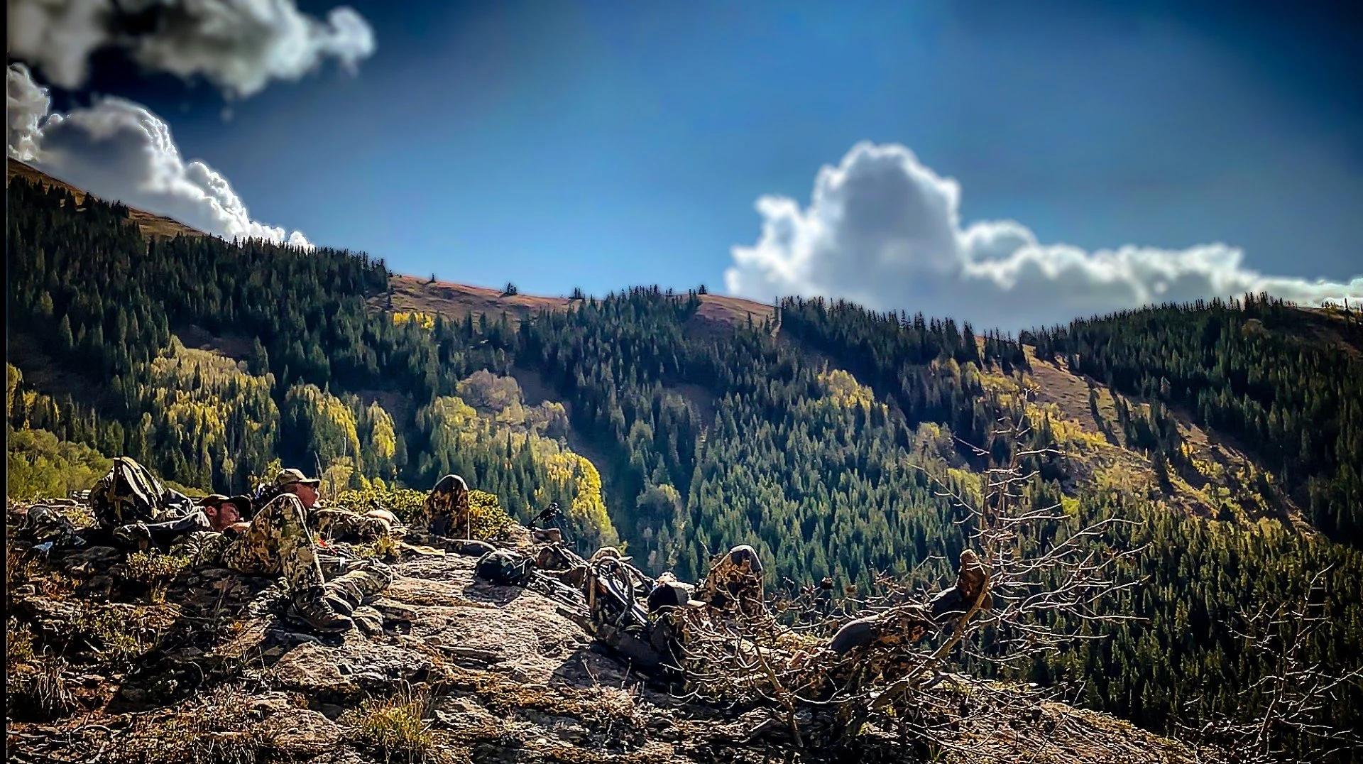 Hikers resting on a rocky trail in a mountainous forested area under a partly cloudy sky.