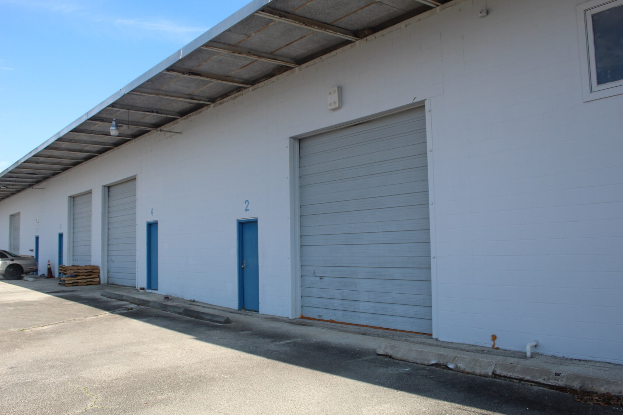 Exterior of a warehouse with multiple garage doors, numbered 2, 4, and others, with a small blue door labeled 2 and a car parked nearby. The building has a white exterior wall and a metal roof extension.