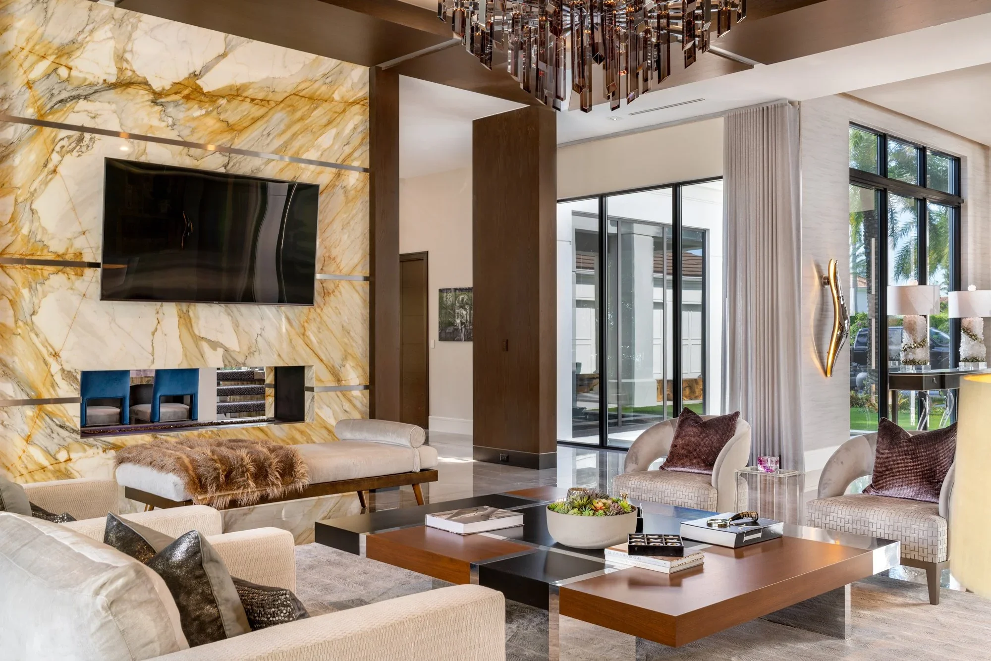 Modern living room with large windows, a marble accent wall with a mounted TV, beige sofas, patterned armchairs, and contemporary decor.