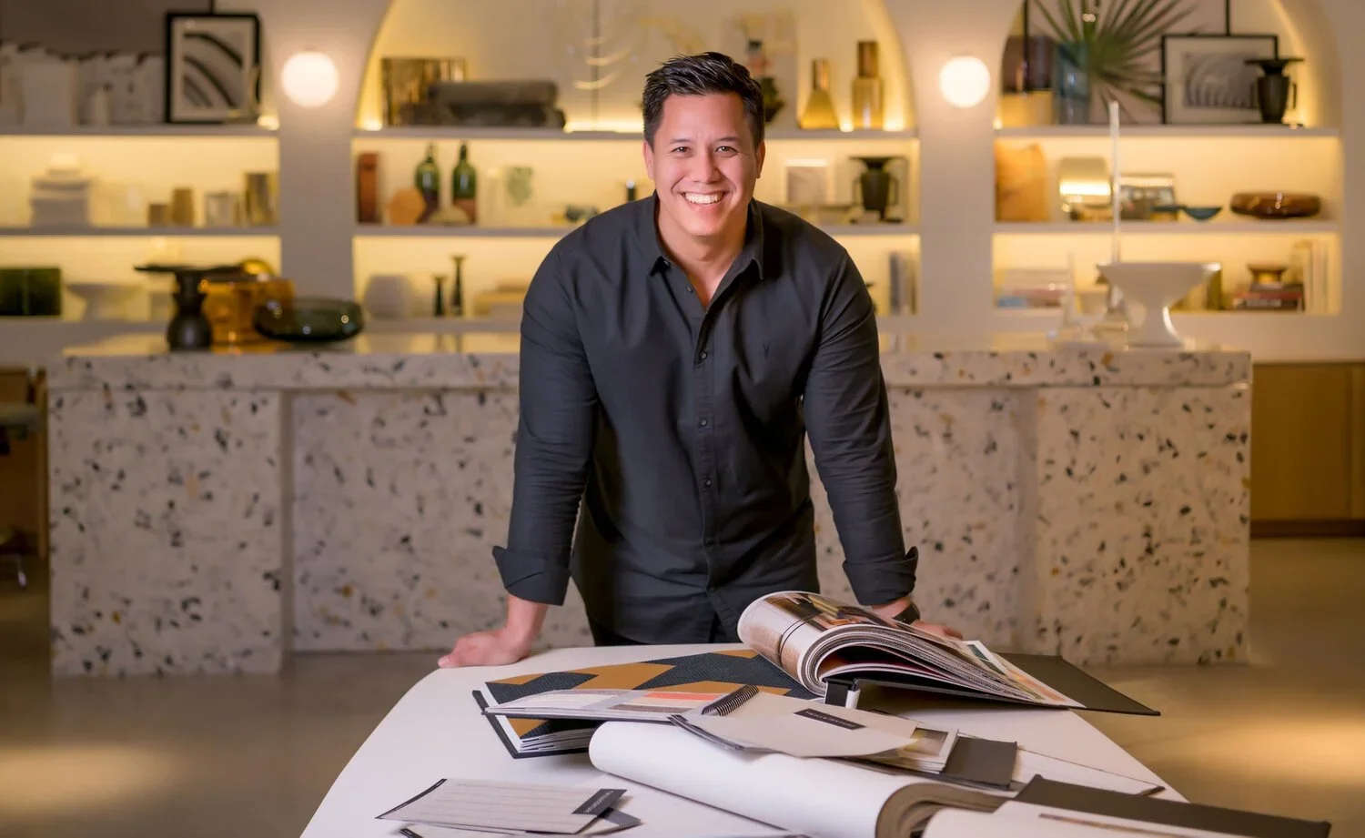A smiling man in a black shirt leaning on a table with open design catalogs in a modern kitchen or showroom with shelves and decorative items in the background.