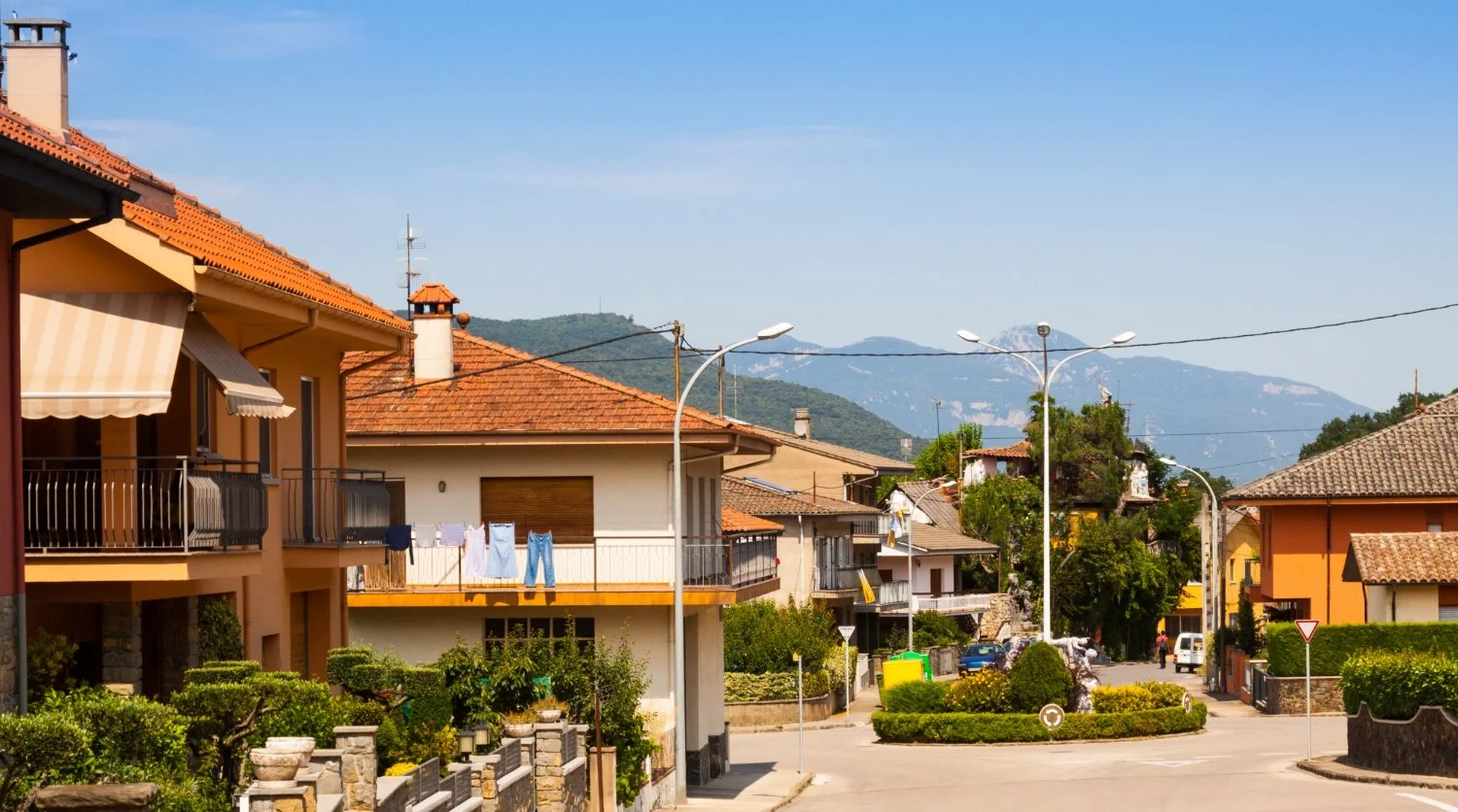 Residential neighborhood with houses, laundry hanging on balconies, green bushes, street lamps, and mountain in the background.