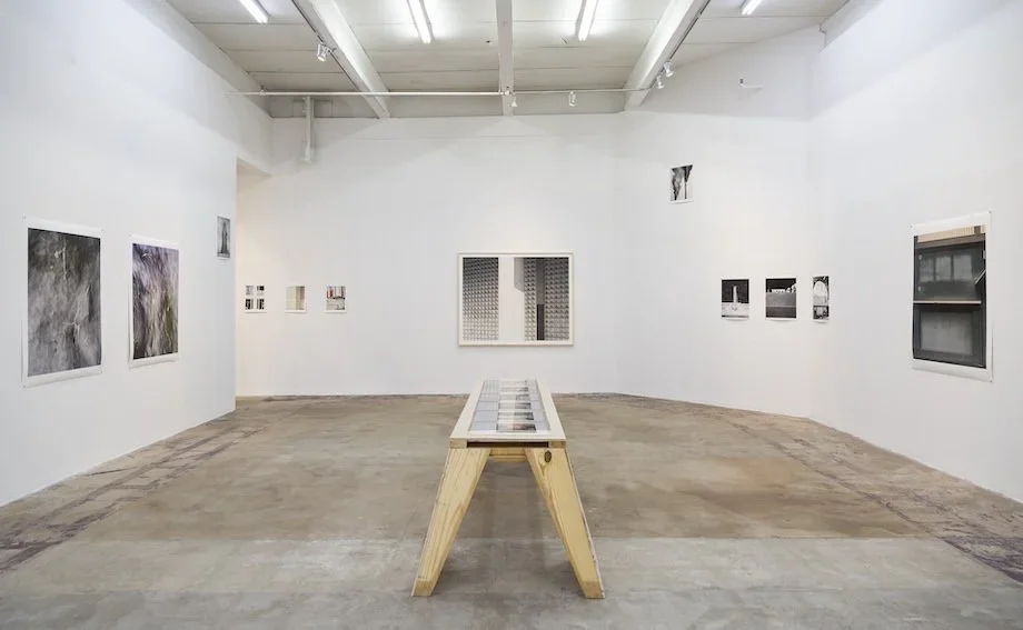 Art gallery with white walls, artwork on display, and a wooden table in the center. The gallery has high ceilings with industrial lighting.