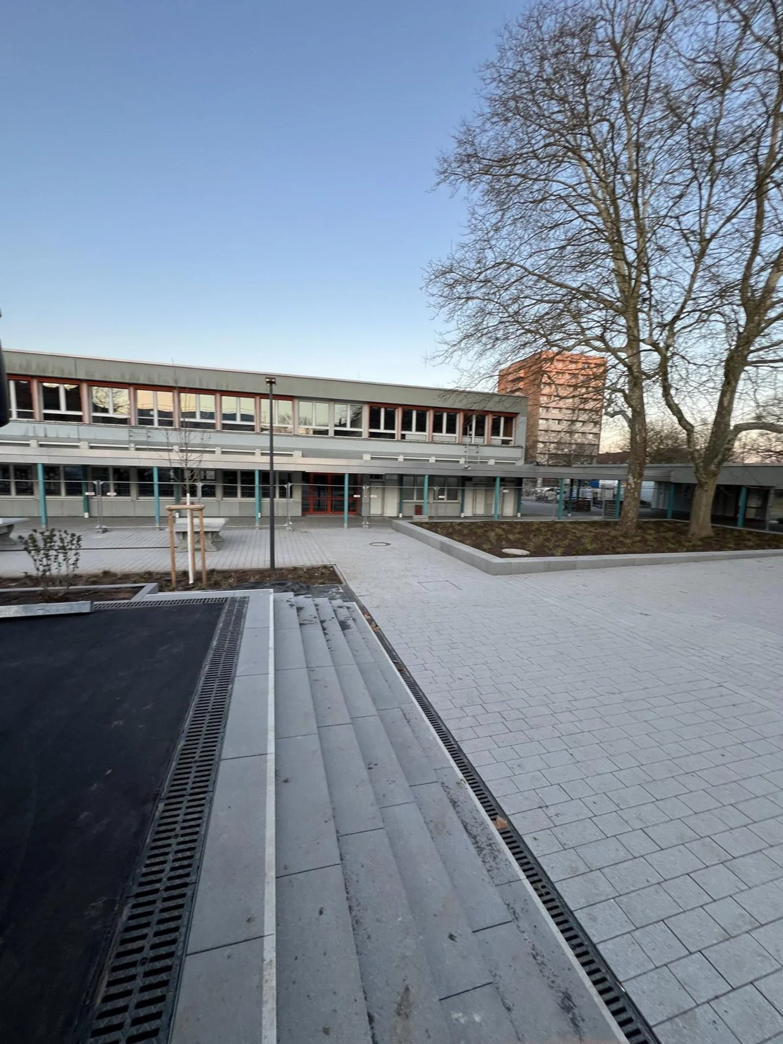 Ein Schulhof mit gepflasterten Wegen, Bäumen und einem mehrstöckigen Schulgebäude im Hintergrund, das bei Sonnenuntergang beleuchtet wird.