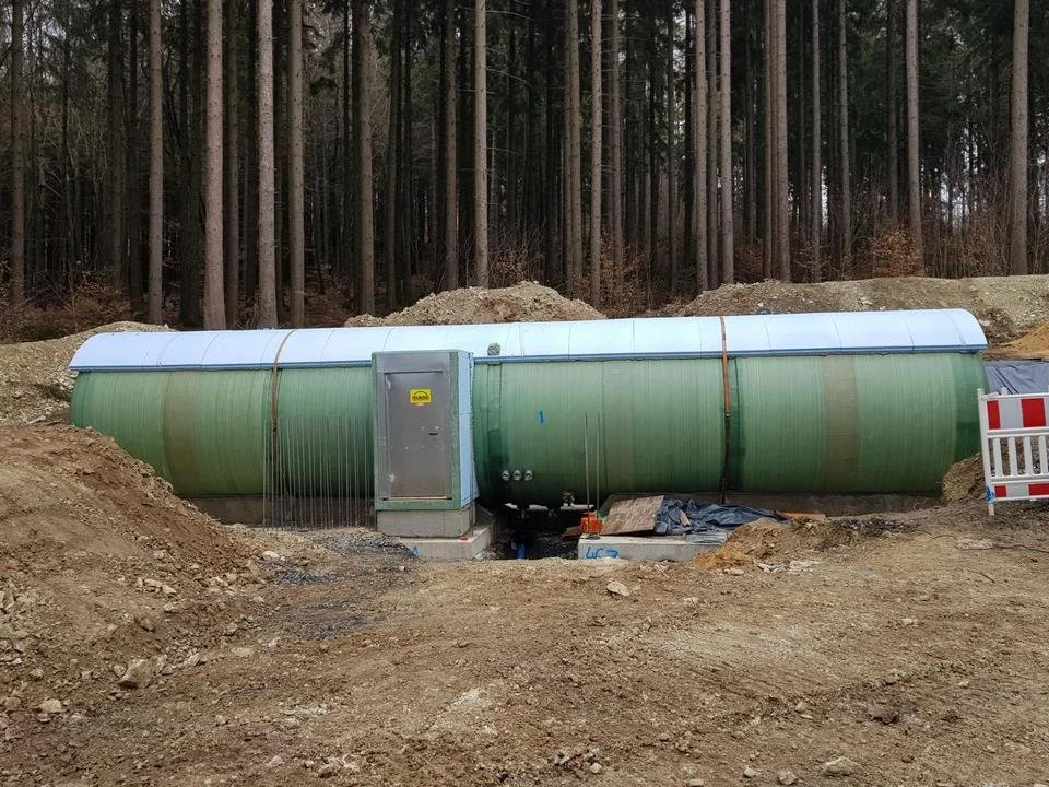 Ein unterirdischer Tankspeicher, umgeben von Erde, vor einem Wald mit hohen Bäumen.