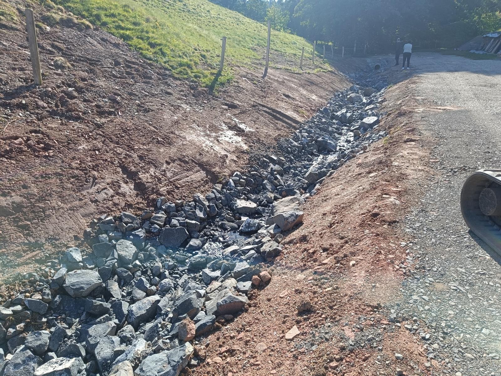 Ein Bauprojekt mit einem ausgehobenen Graben neben einem unbefestigten Weg, auf dem einige Menschen arbeiten oder spazieren gehen.