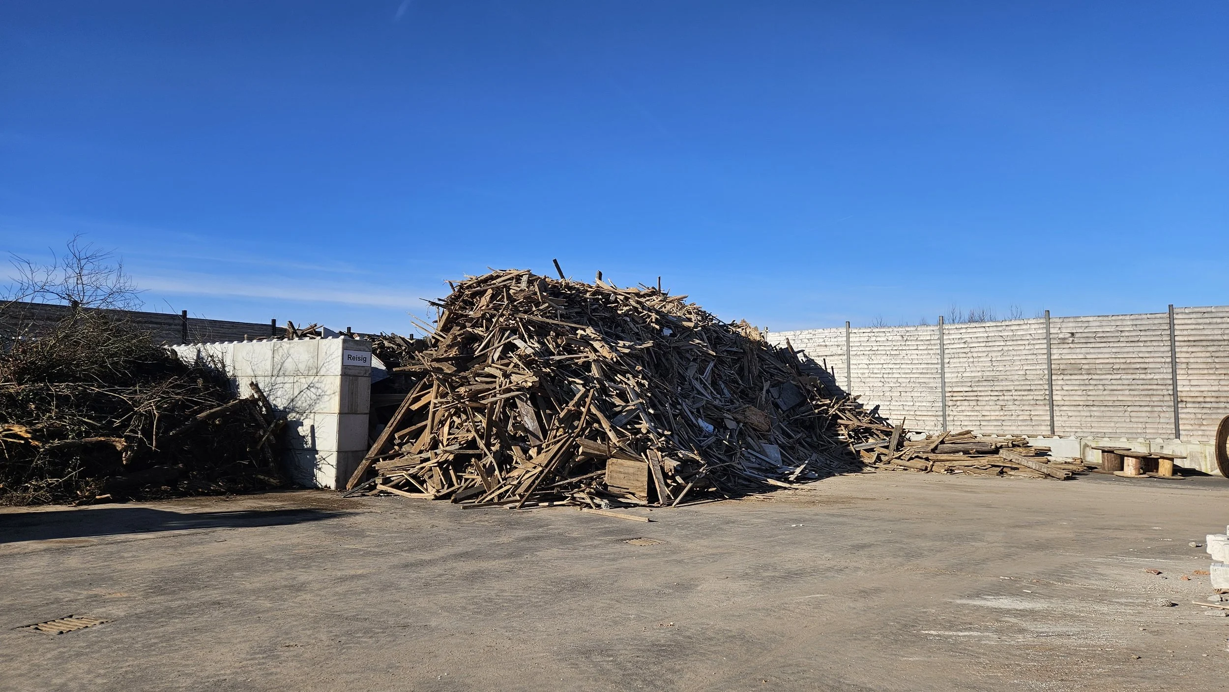 Haufen mit Holzreste und Sperrmüll vor einer weißen Wand und einem hohen Zaun bei sonnigem Himmel.